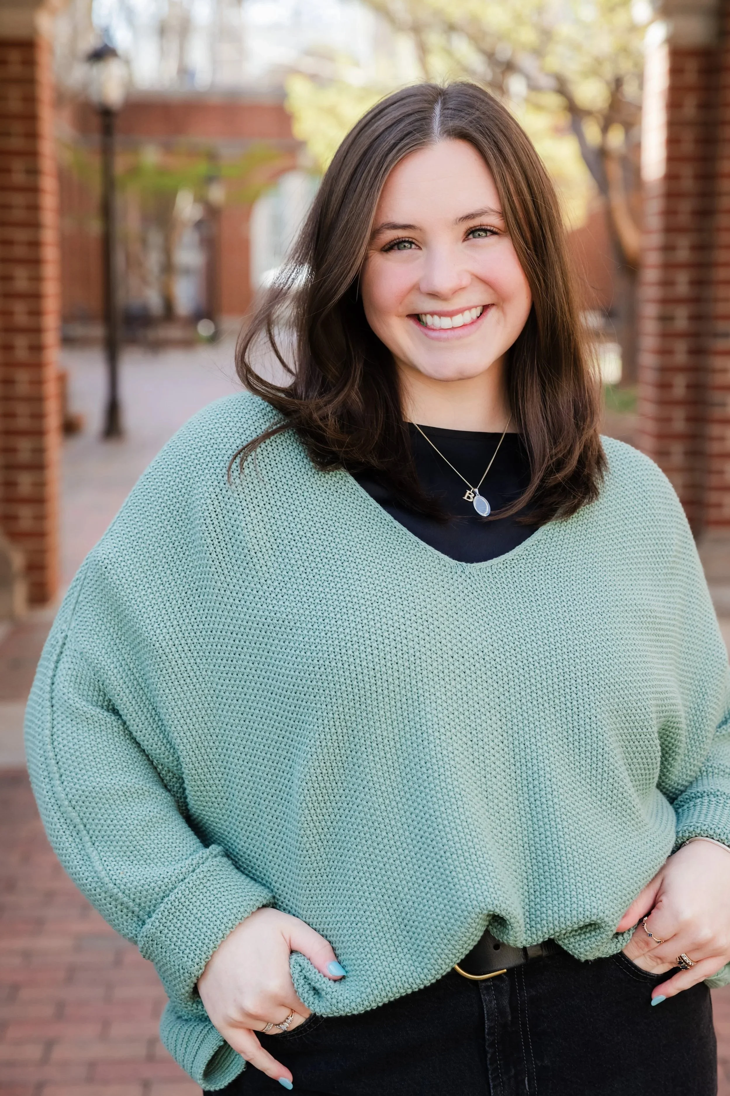 Close-up of a young woman with long brown hair smiling outdoors during daytime, wearing a mint green sweater over a black top, with trees and brick structures in the background.