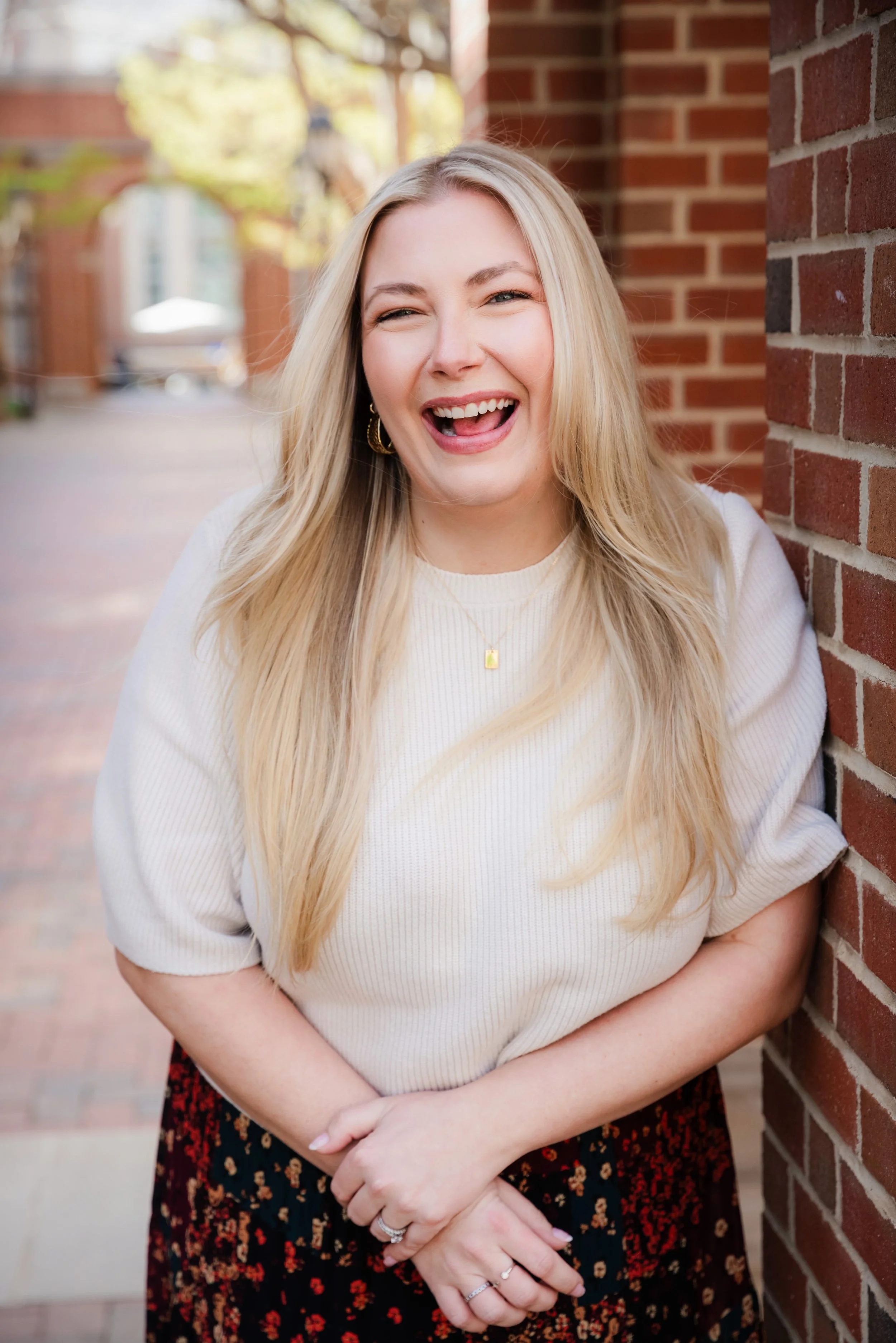 A smiling woman with long blonde hair, wearing a white top and a floral skirt, standing outdoors against a brick wall and a blurred background of trees and buildings.