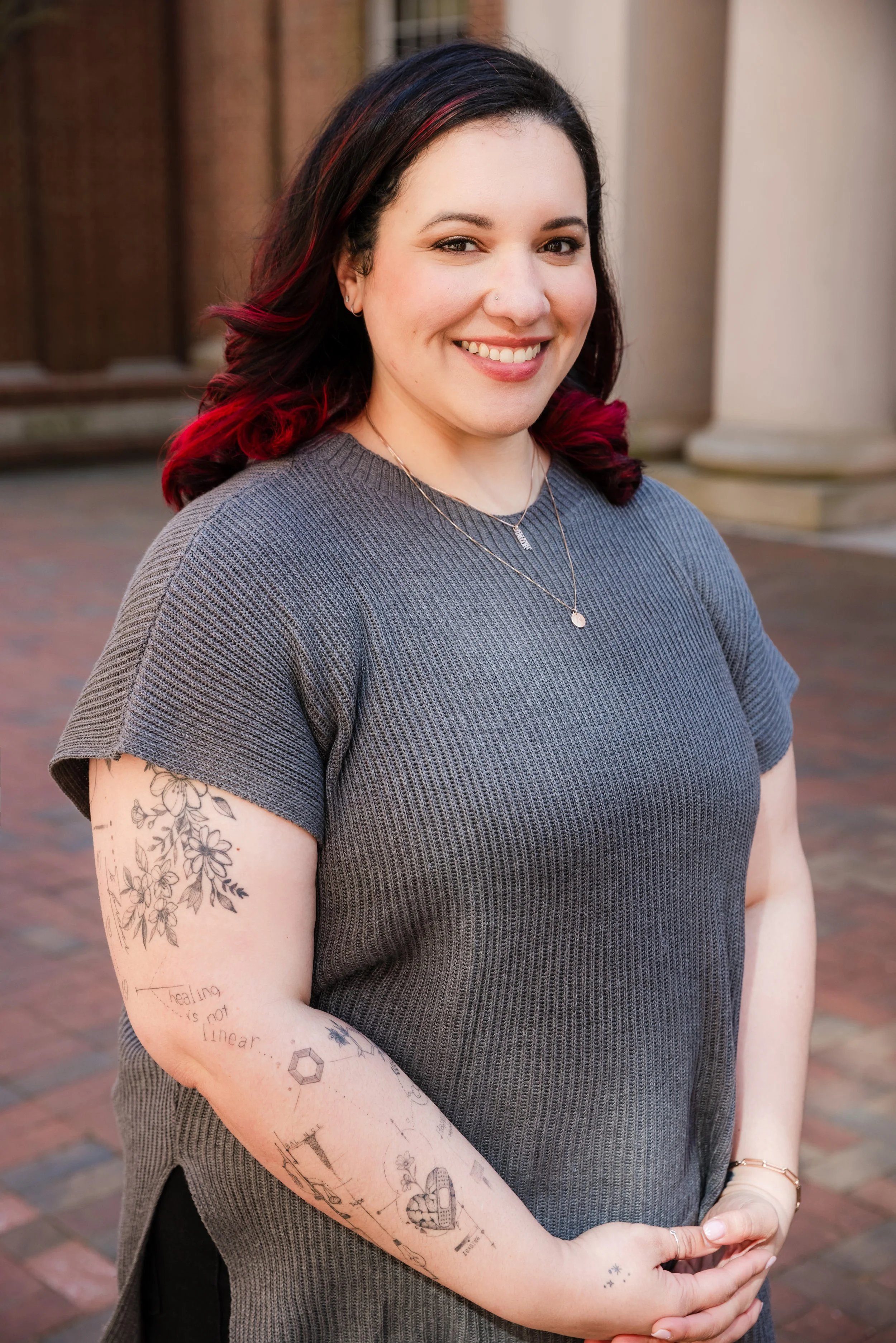 An approachable looking therapist smiling outdoors, wearing a gray short-sleeve top, with dark hair with red highlights, and showing tattoos on her arms.