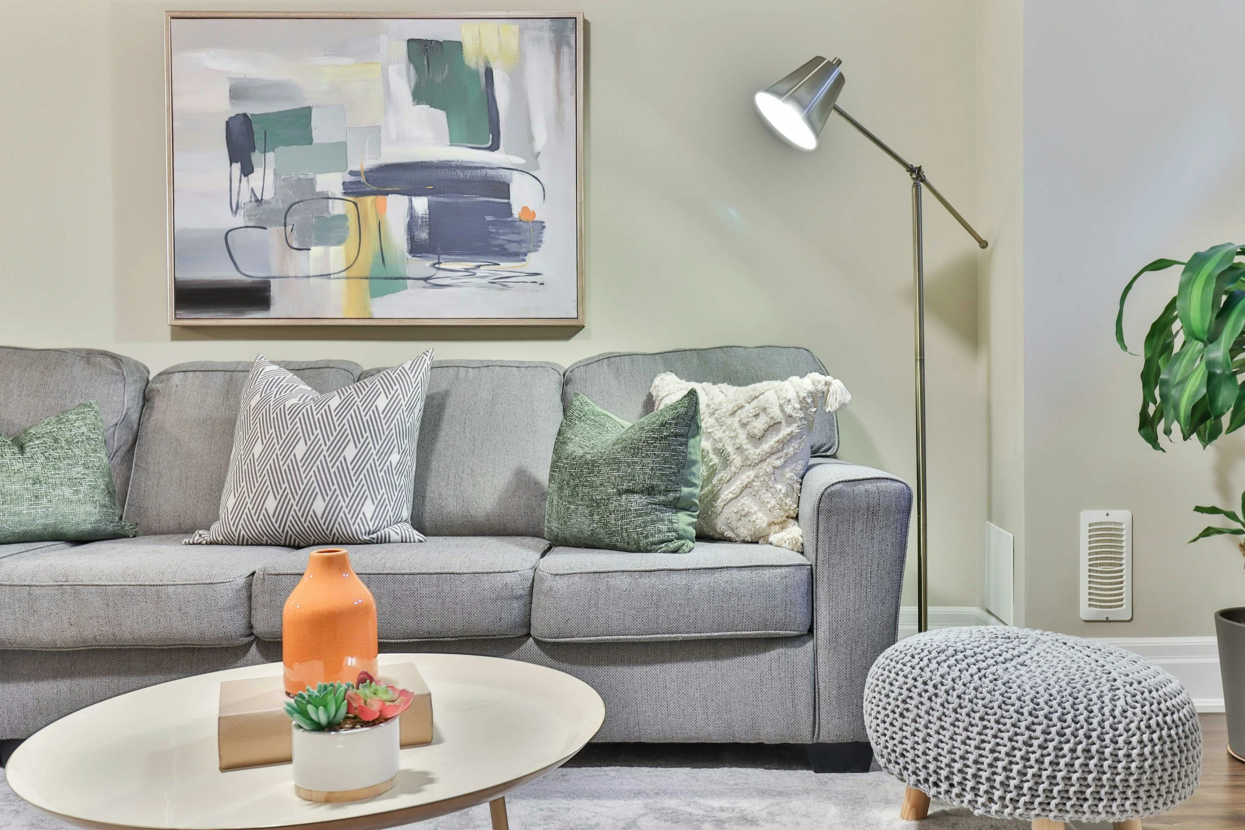 Airbnb example - Living room with a gray sofa, decorative pillows, an abstract painting on the wall, a tall floor lamp, a potted plant, a white coffee table with a vase and succulent, and a knit pouf.