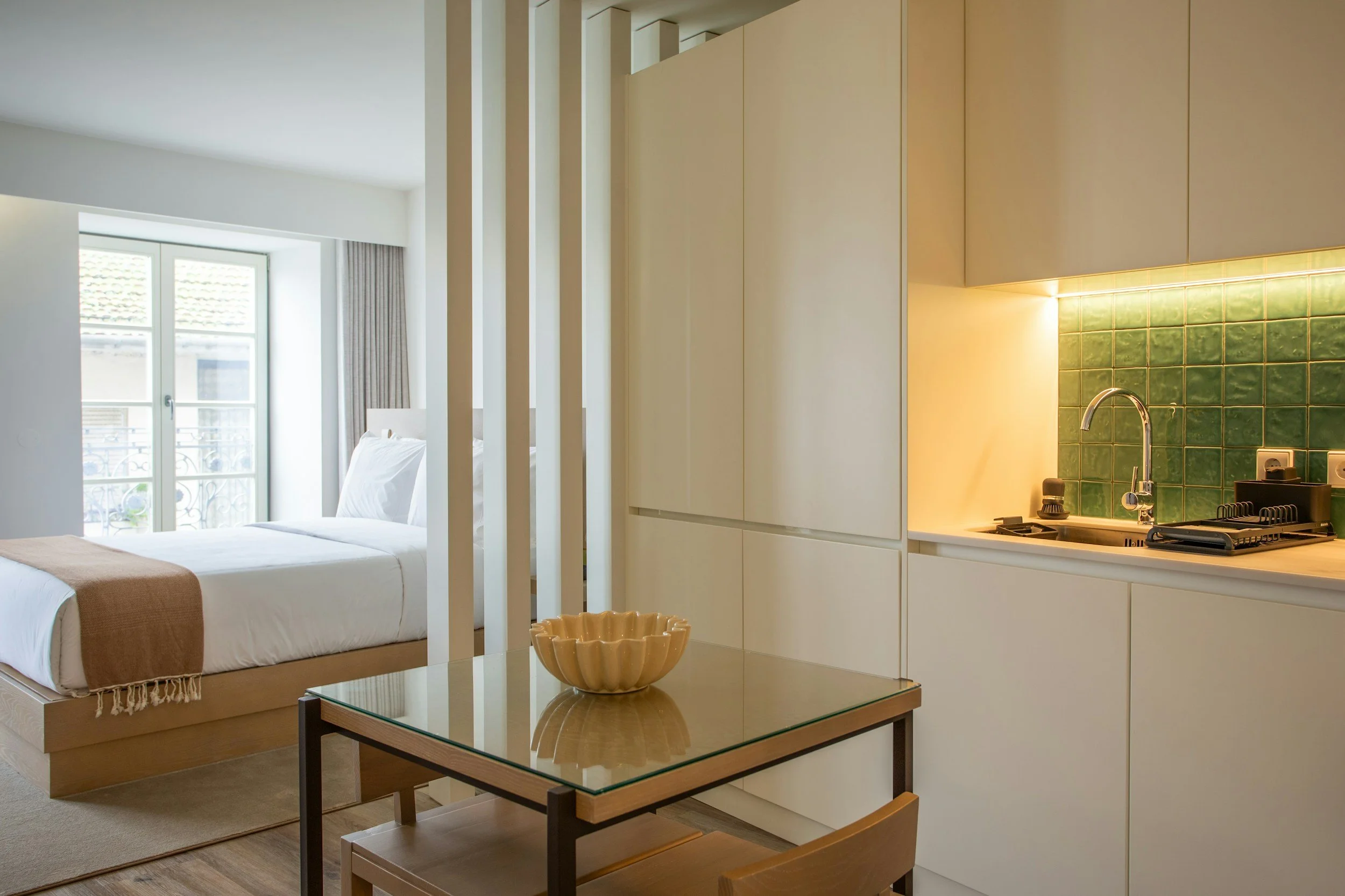 Airbnb example - A studio apartment with a bed near a large window, a small dining table with a ceramic bowl, and a kitchenette with green tiled backsplash and white cabinets.