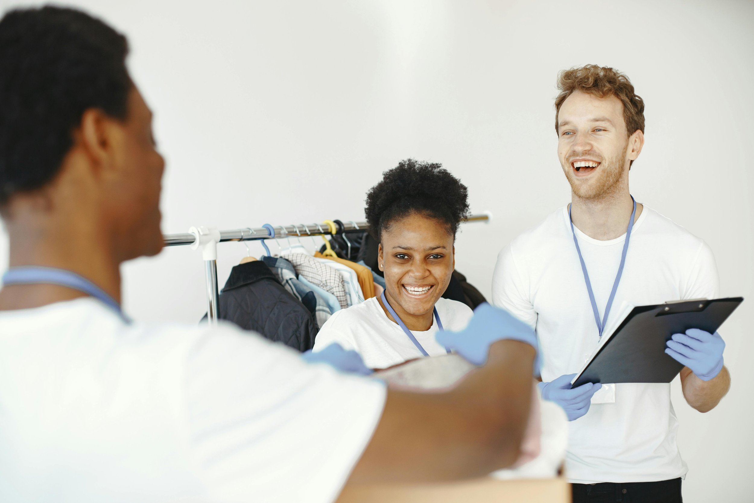 Three volunteers in white shirts with blue gloves and ID badges share a joyful moment in a clothing donation center, with clothes on a rack behind them.