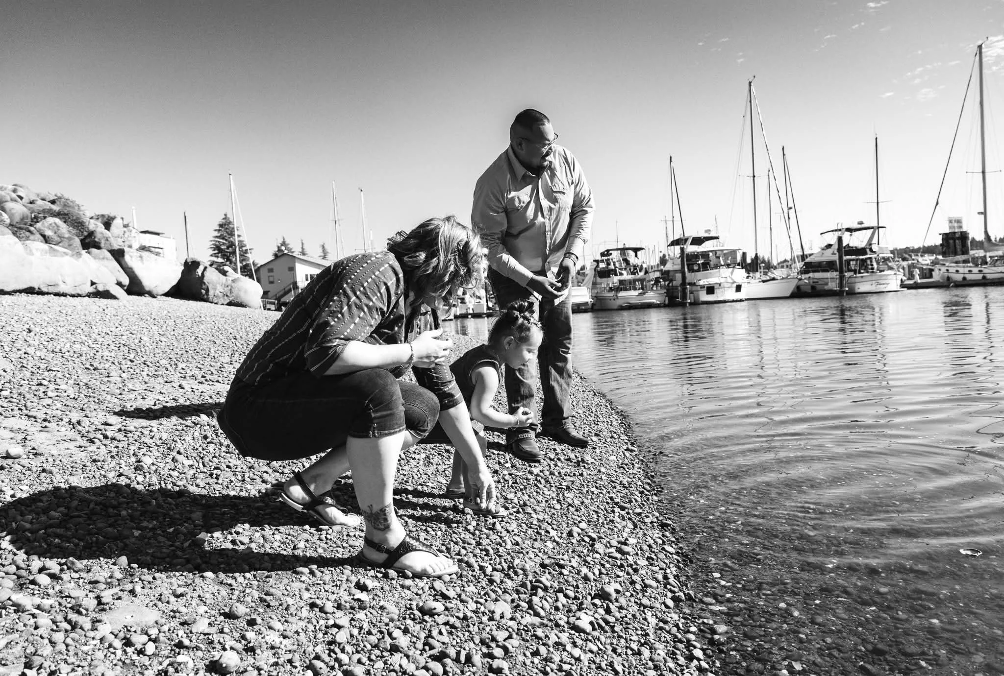 A family of three, two adults and a child, crouching by the water's edge at a marina, with sailboats and yachts docked nearby, on a sunny day.
