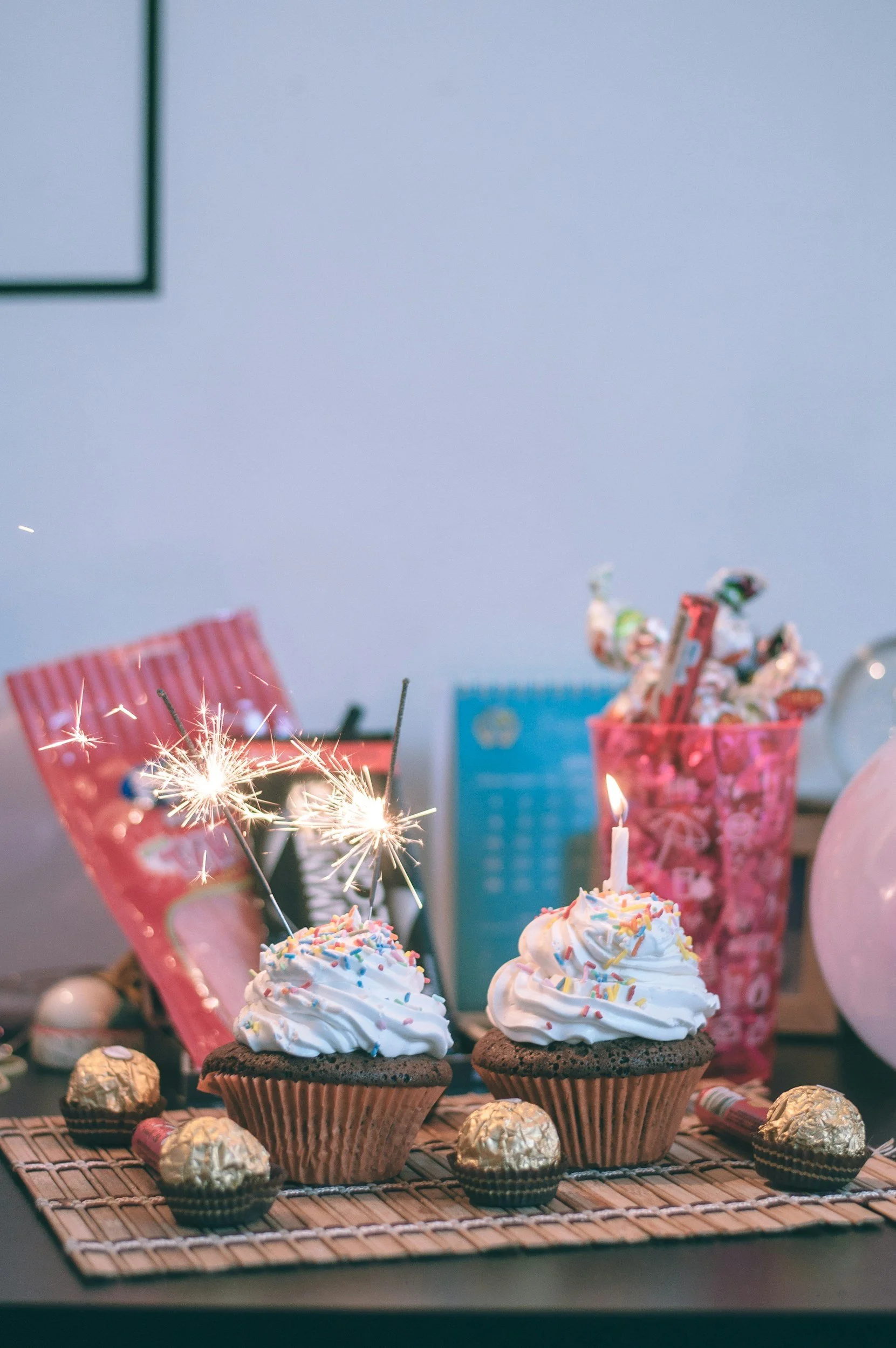 Celebration table with cupcakes, chocolates, and sparklers, pink and blue party decor, and candy in a pink cup.