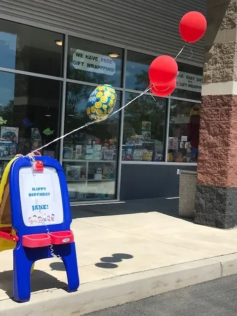 Colorful balloons tied to a small blue and red plastic easel outside a store, with a birthday message for Jake on the easel, and signs in the window offering free gift wrapping.