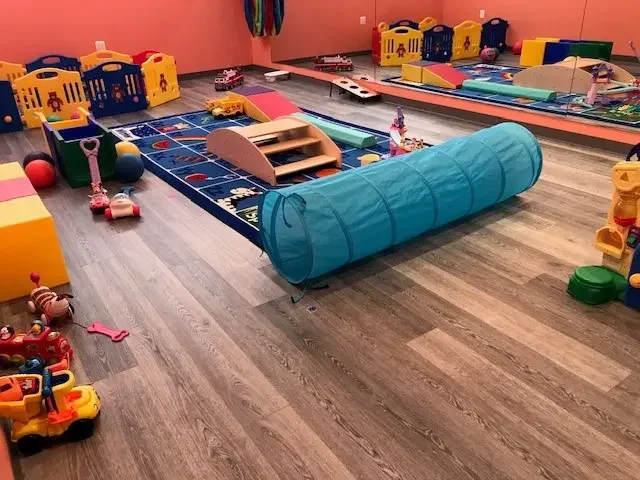 Indoor playroom with colorful toys, a blue tunnel, a small balancing bridge, and a train-themed rug on a wooden floor.