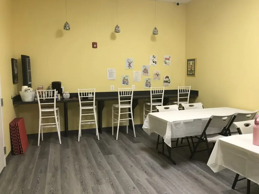 A small room with yellow walls and gray wood flooring, featuring high chairs at a black counter, and tables with white tablecloths and folding chairs, some with items on them, with framed artwork and colorful drawings on the walls.
