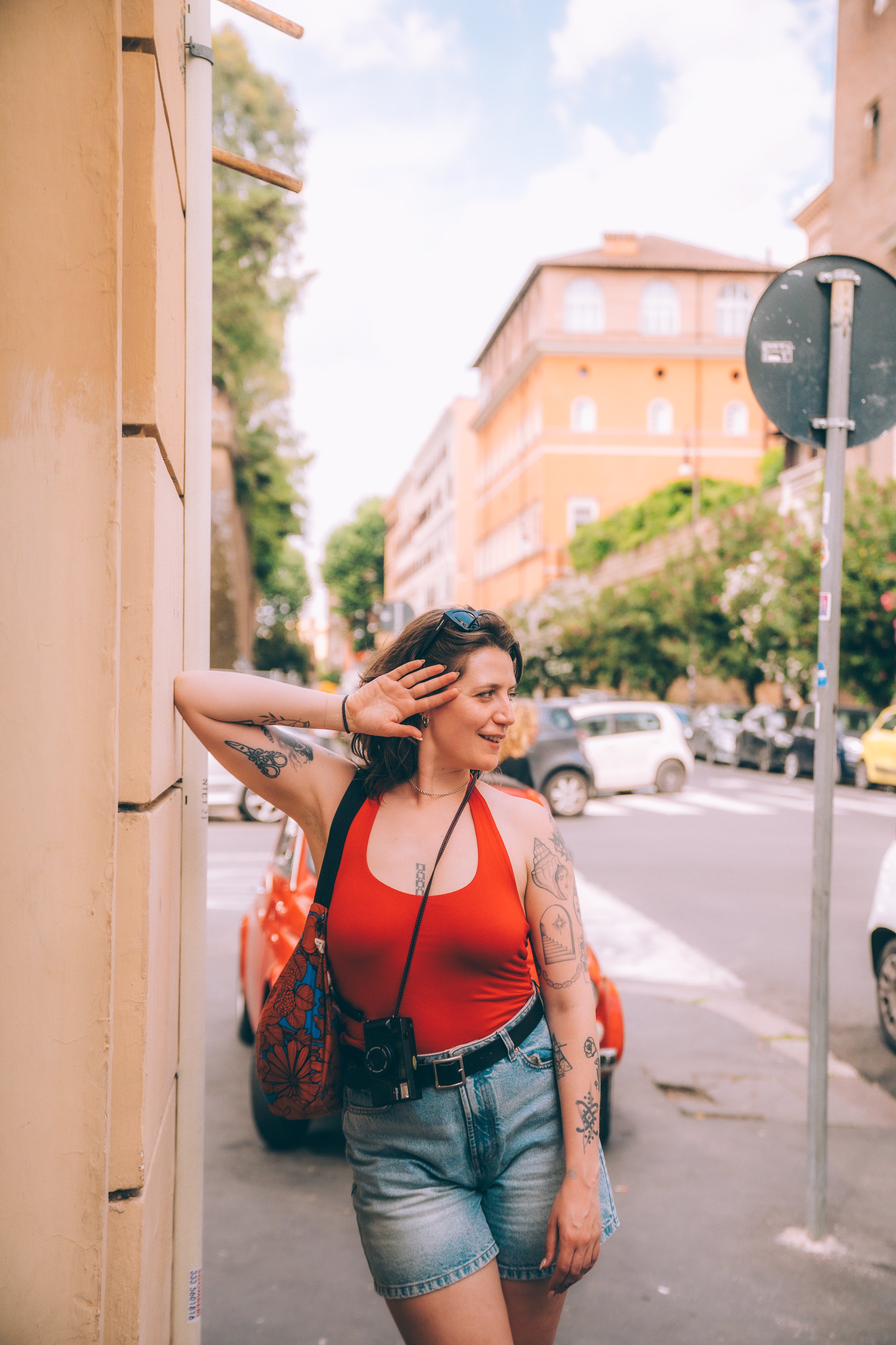 A woman with tattoos on her arms and a camera hanging around her neck, leaning against a yellow building, smiling and shielding her eyes from the sun on a city street with parked cars and buildings in the background.