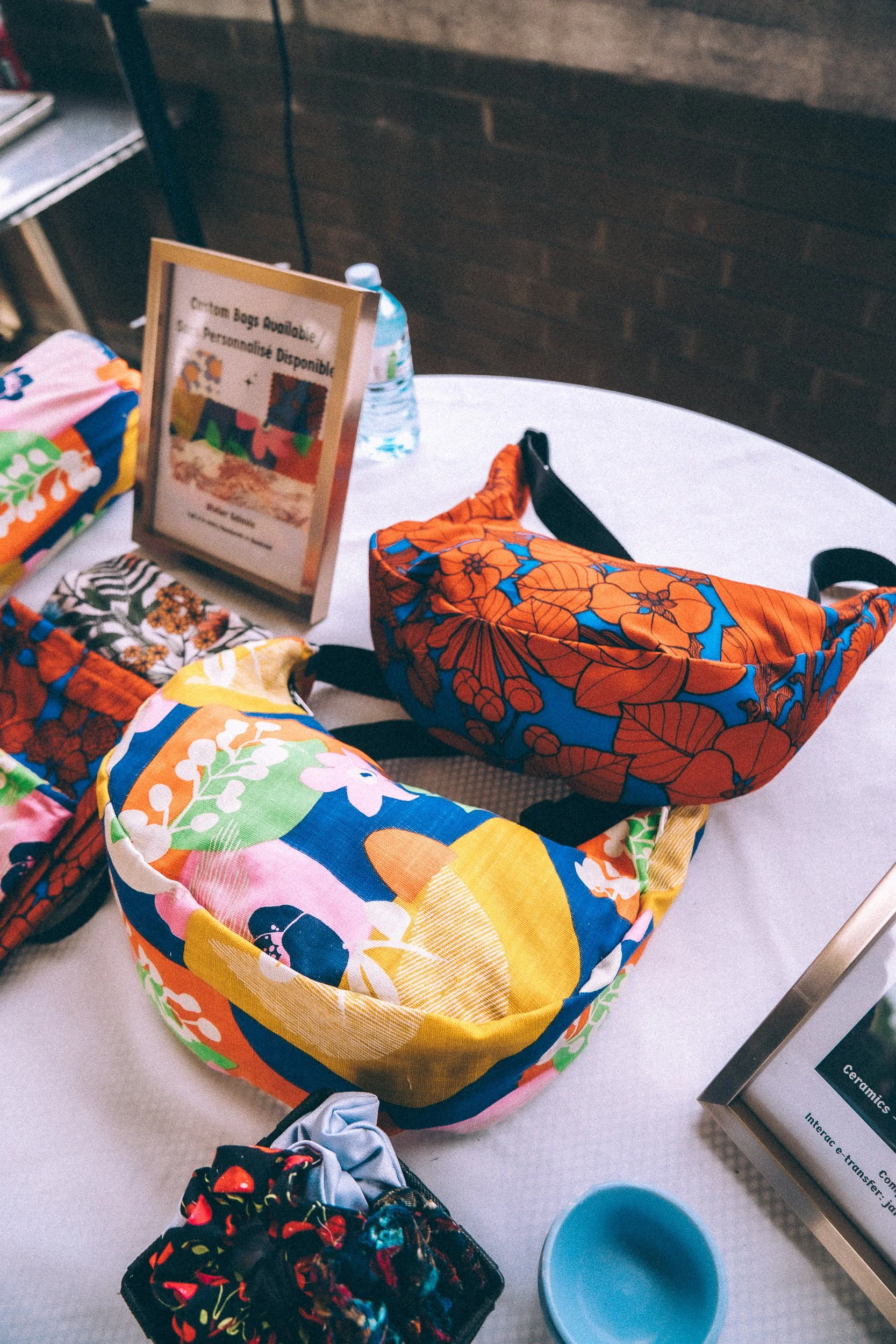 Colorful fabric bags with floral patterns on a table, with a water bottle and a framed sign in the background.