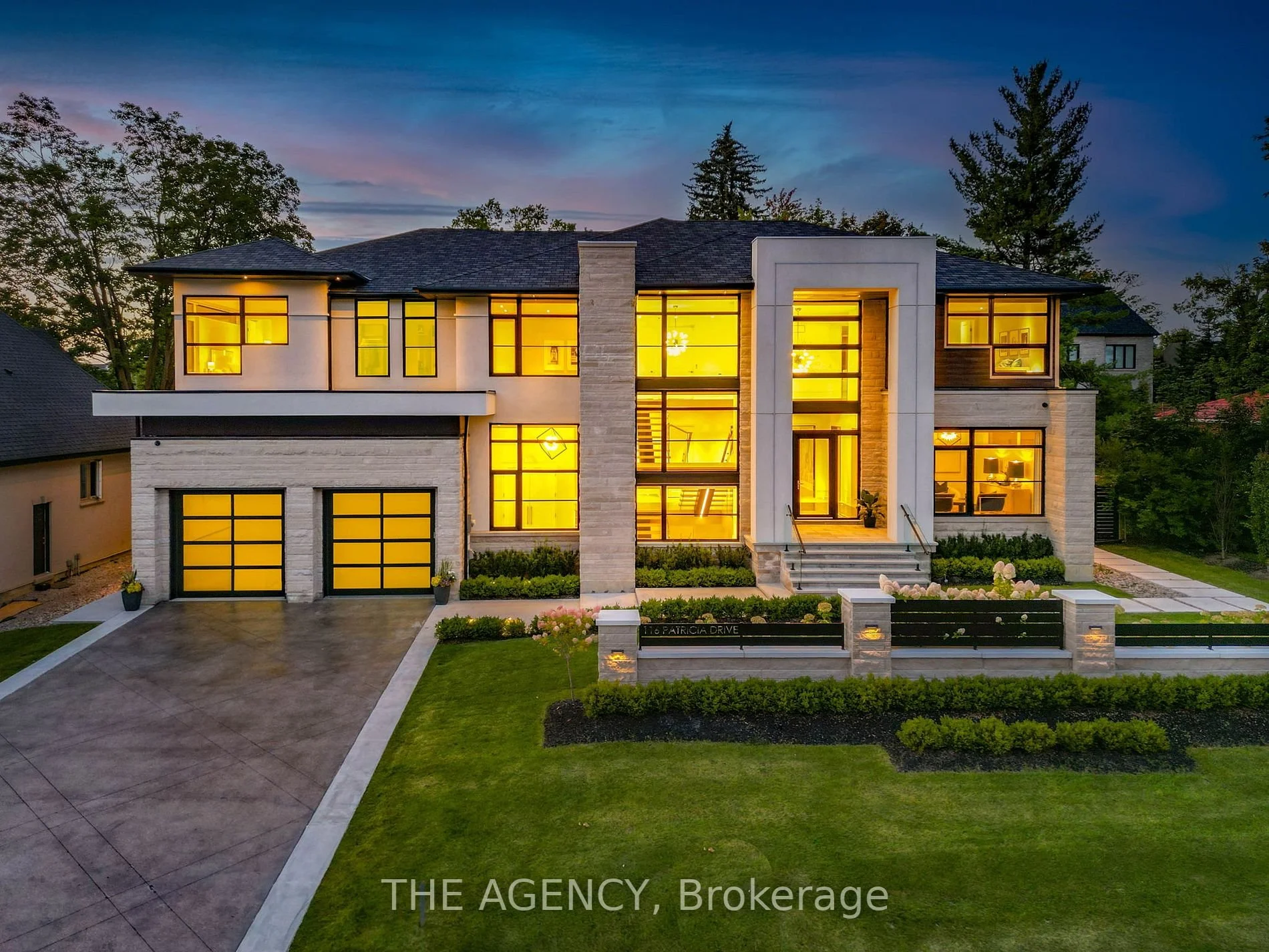 Modern two-story house with large windows illuminated from inside, a driveway leading to a two-car garage, manicured lawn, and landscaped garden, at dusk.
