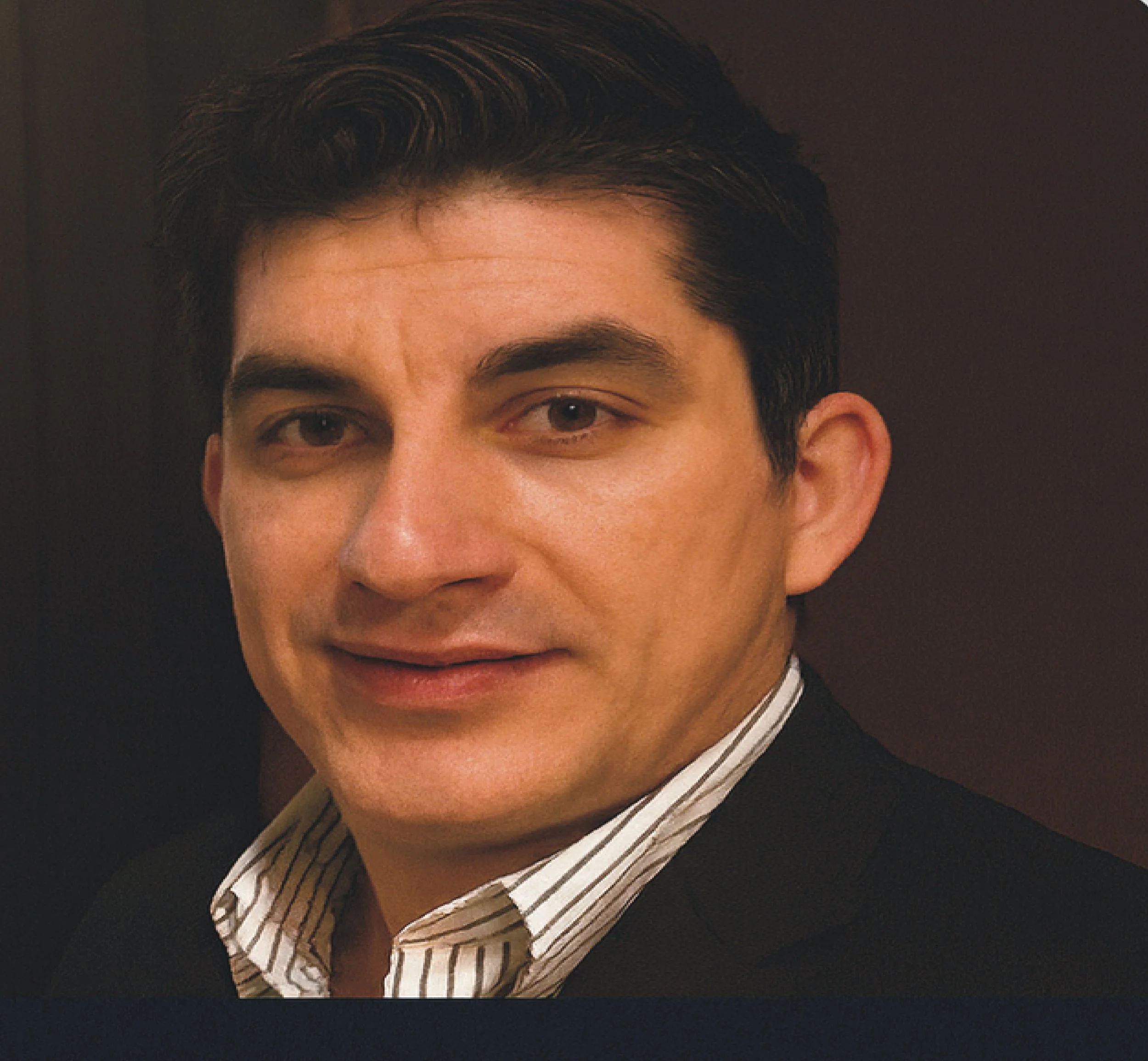 Close-up of a man with dark hair, wearing a striped dress shirt and black blazer, looking at the camera with a neutral expression.