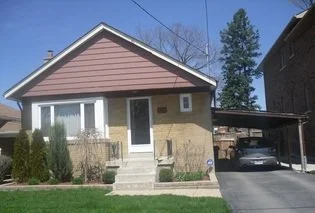 A single-story house with beige and brown exterior, front lawn, and a driveway with a carport on the right.