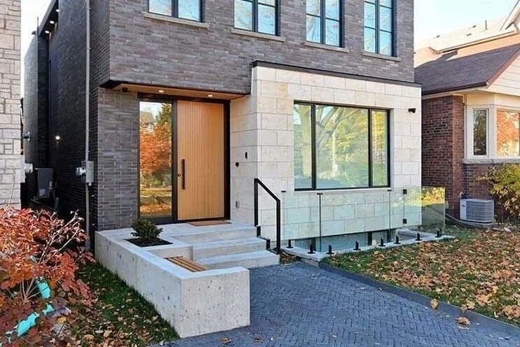 Modern house exterior with a combination of dark brick and white stone facade, a wooden front door, large windows, black metal railing on steps leading to the door, and a small landscaped area with fallen leaves and a gravel driveway.