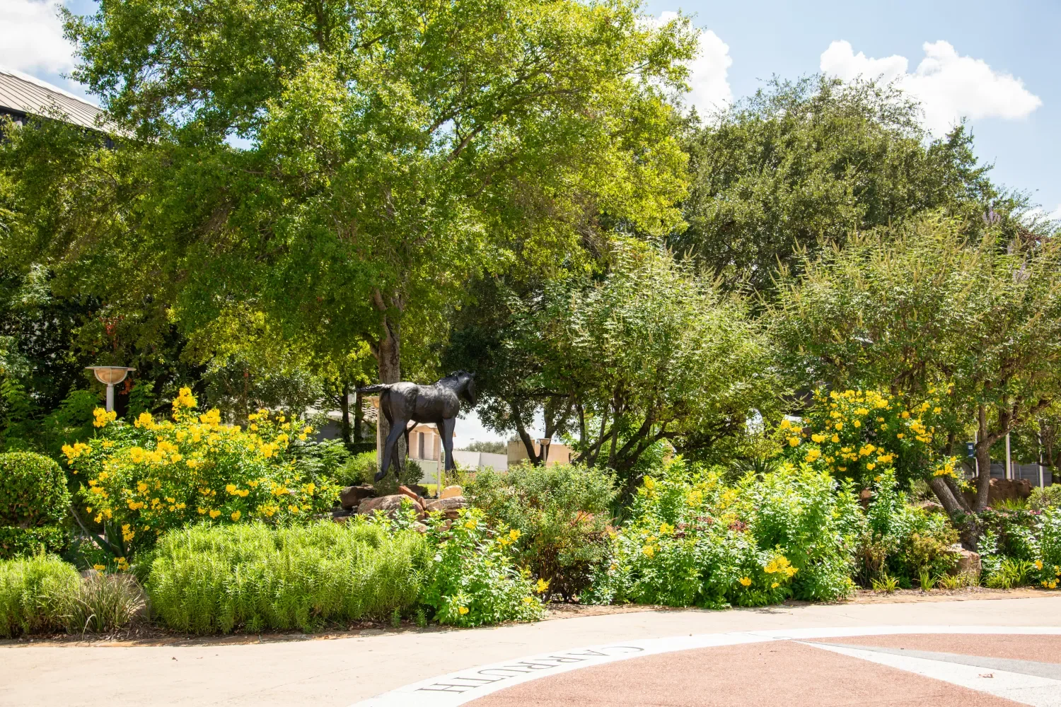 A lush green garden with trees, yellow flowers, a statue of a horse, and a paved pathway under a partly cloudy sky.