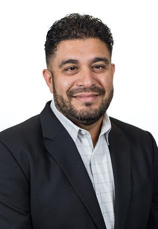 Portrait of a man with dark, curly hair and a beard, wearing a black blazer and a collared shirt, smiling against a white background.