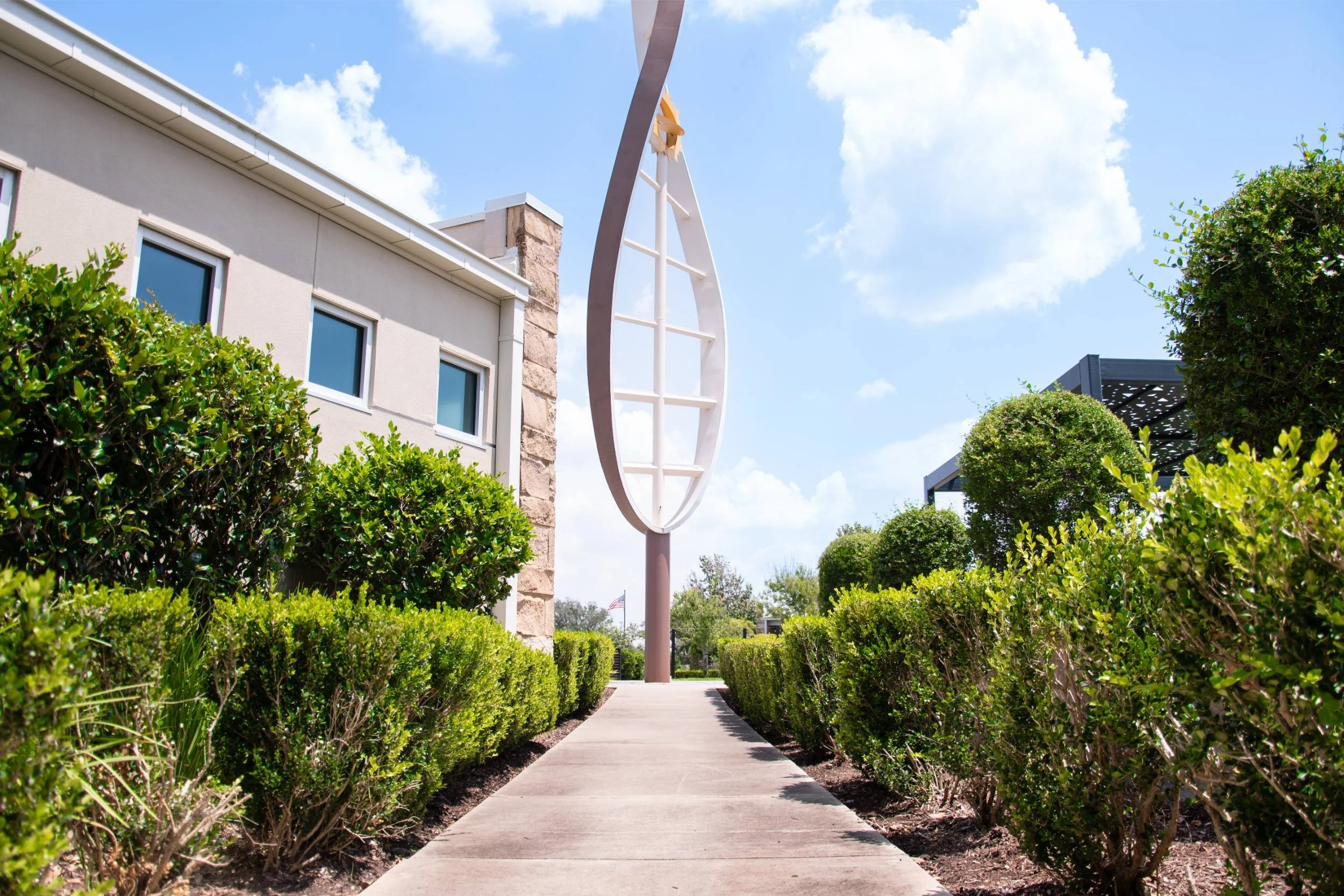 A sidewalk leading to a tall modern sculpture outdoors, with bushes and plants on both sides, under a blue sky with scattered clouds in Houston, TX 
