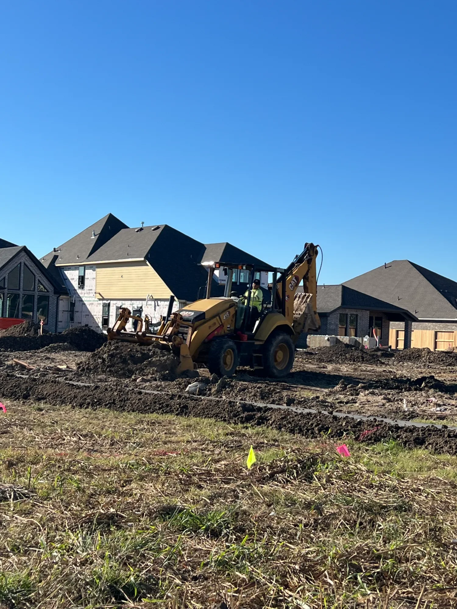 Construction site with a yellow bulldozer operating on a dirt lot in front of residential houses under a clear blue sky.