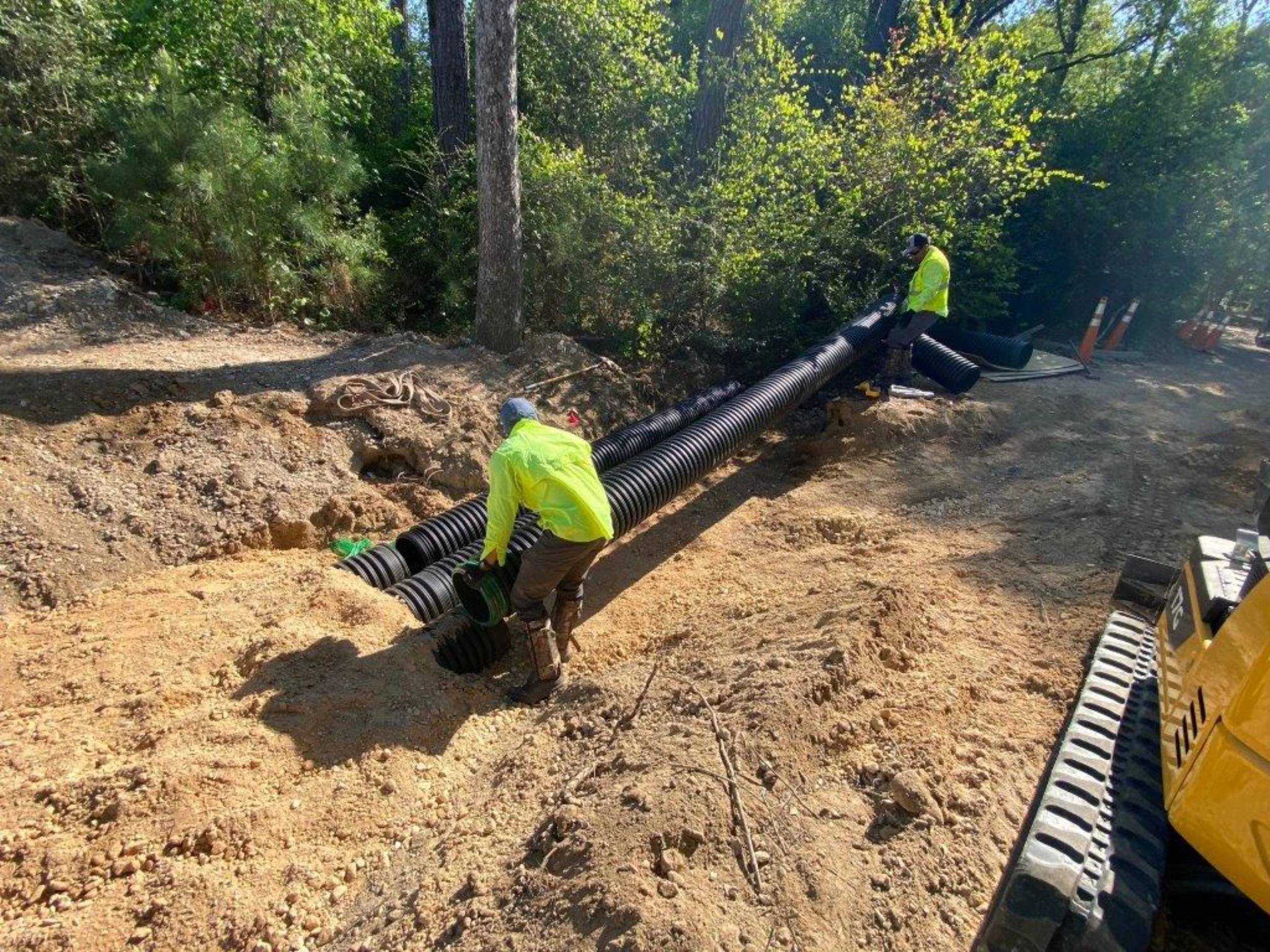 Commercial landscaping workers in yellow safety vests install large black pipe underground in a dirt road surrounded by trees in Katy, TX