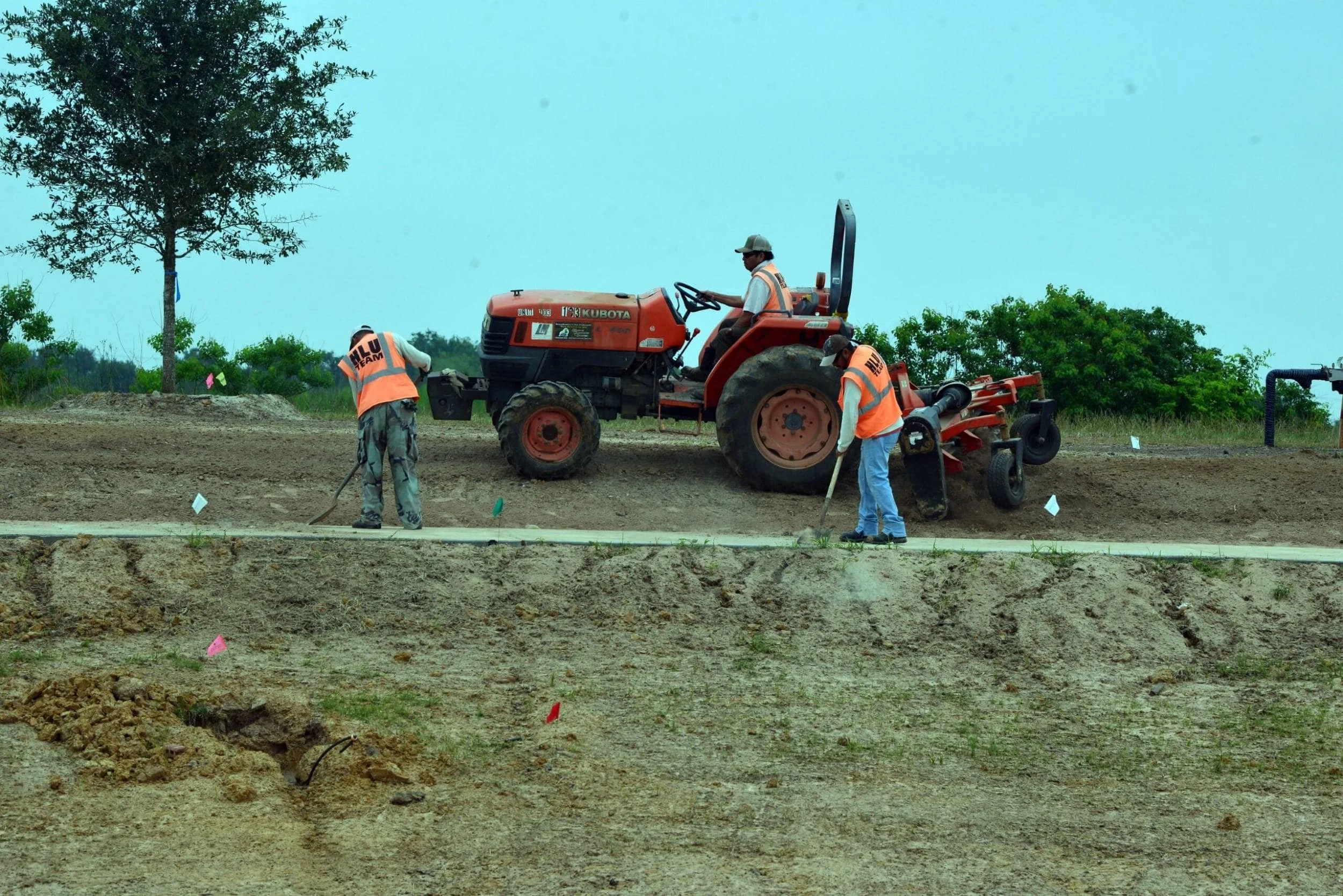 Commercial landscaping company  workers working on a dirt construction site with a red tractor in the background, trees, and a cloudy sky in Houston, TX
