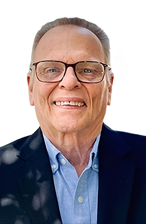 An older man with glasses smiling, wearing a blue shirt and a dark blazer, against a plain white background.
