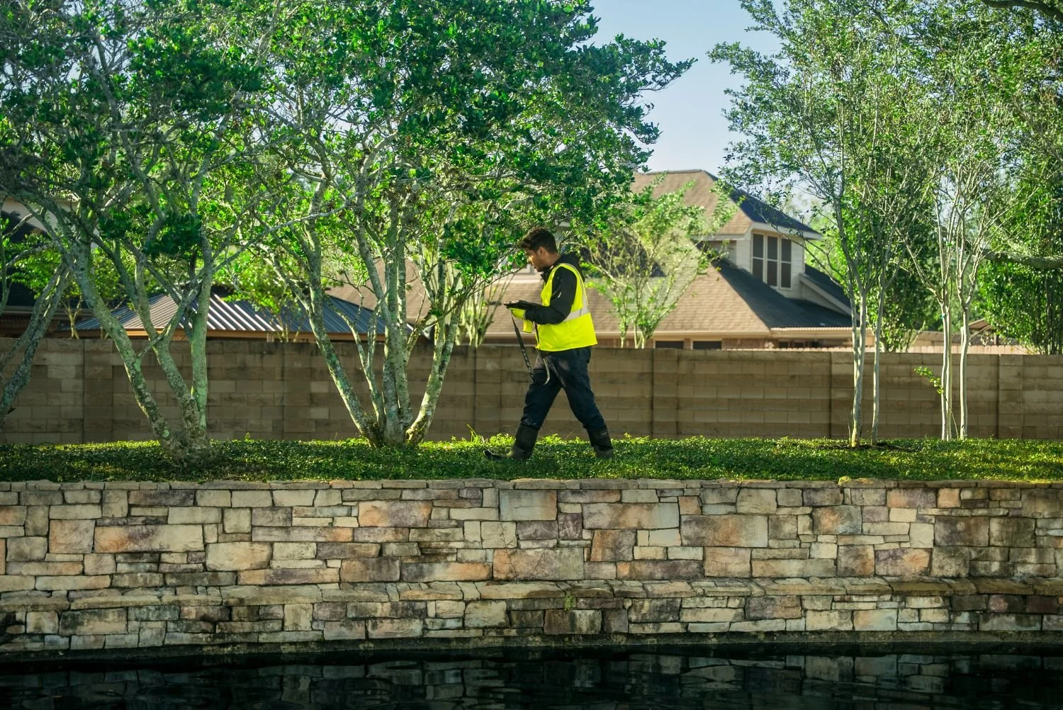 landscape technician in safety vest checking tree line beside stacked stone wall as part of a commercial landscaping company service in Chambers County, TX