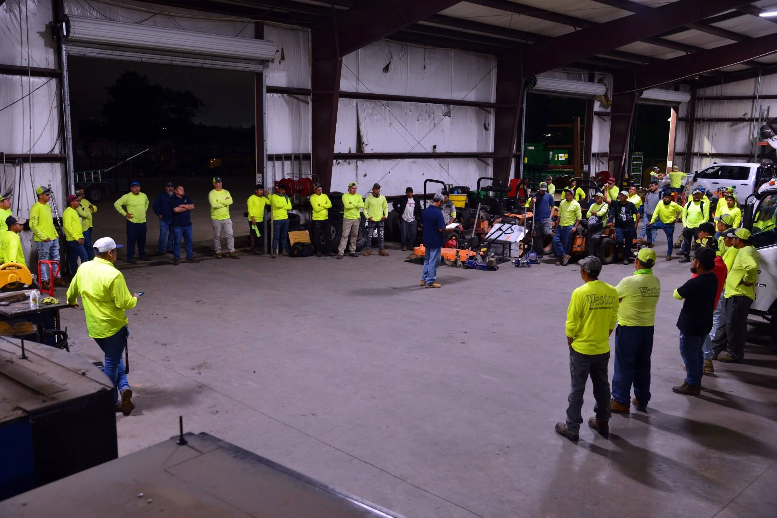 A group of workers wearing high-visibility yellow shirts stand inside a large industrial warehouse, listening to a person speaking at the center. The warehouse has open doors, revealing an outdoor area Harris County, TX 
