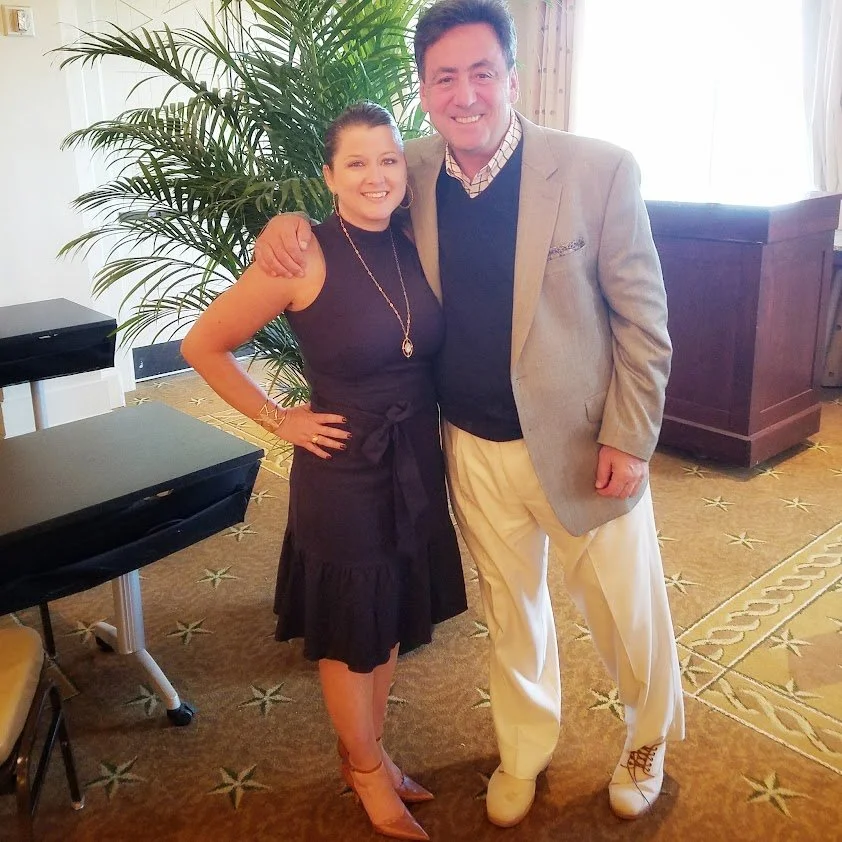 A man and woman standing together indoors, smiling at the camera. The woman is wearing a sleeveless black dress with a necklace, while the man is dressed in a light gray blazer, dark sweater, and cream-colored pants. They are standing in front of a large green plant, with a dark wooden piece of furniture and light curtains in the background.