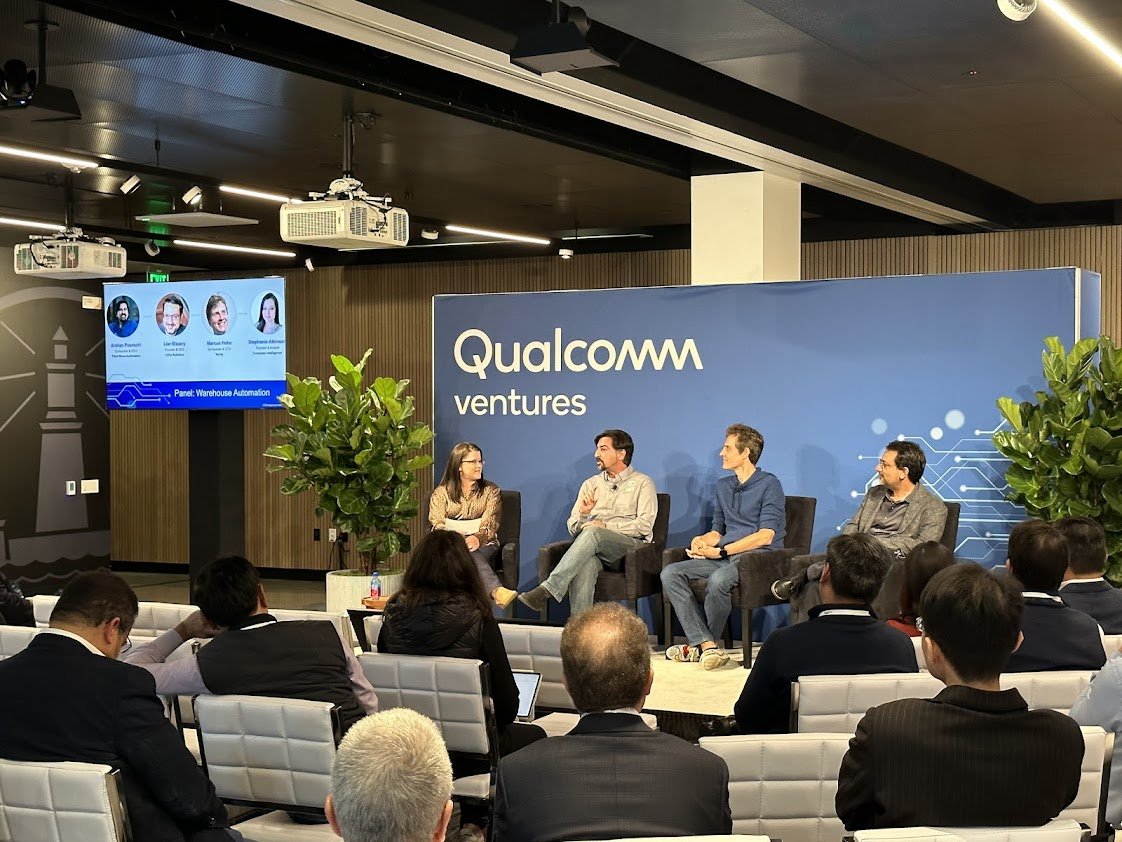 Panel discussion at Qualcomm Ventures event with four speakers seated on a stage, audience seated in front, large Qualcomm Ventures banner and a digital screen displaying panel information.