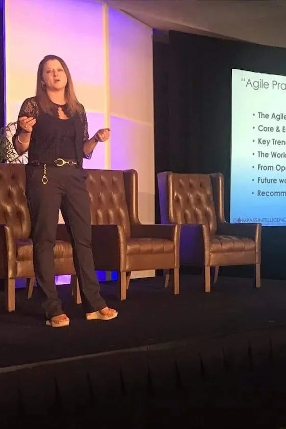 Stephanie Atkinson, founder of Compass Intelligence, giving a presentation on a stage on the topic of "Agile Enterprises" and three empty brown chairs behind her.