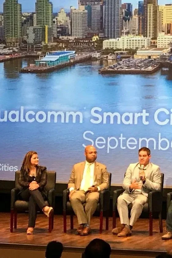 Three people sitting on stage in front of a large screen displaying a city skyline and water. The screen has text reading 'Qualcomm Smart City September'.