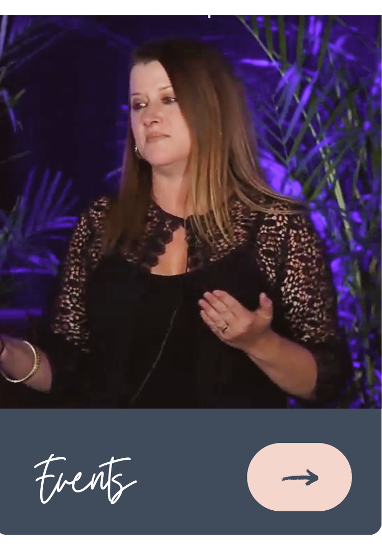 A woman with brown hair wearing a black lace top, holding a black object, in front of a purple-lit backdrop with green leaves.