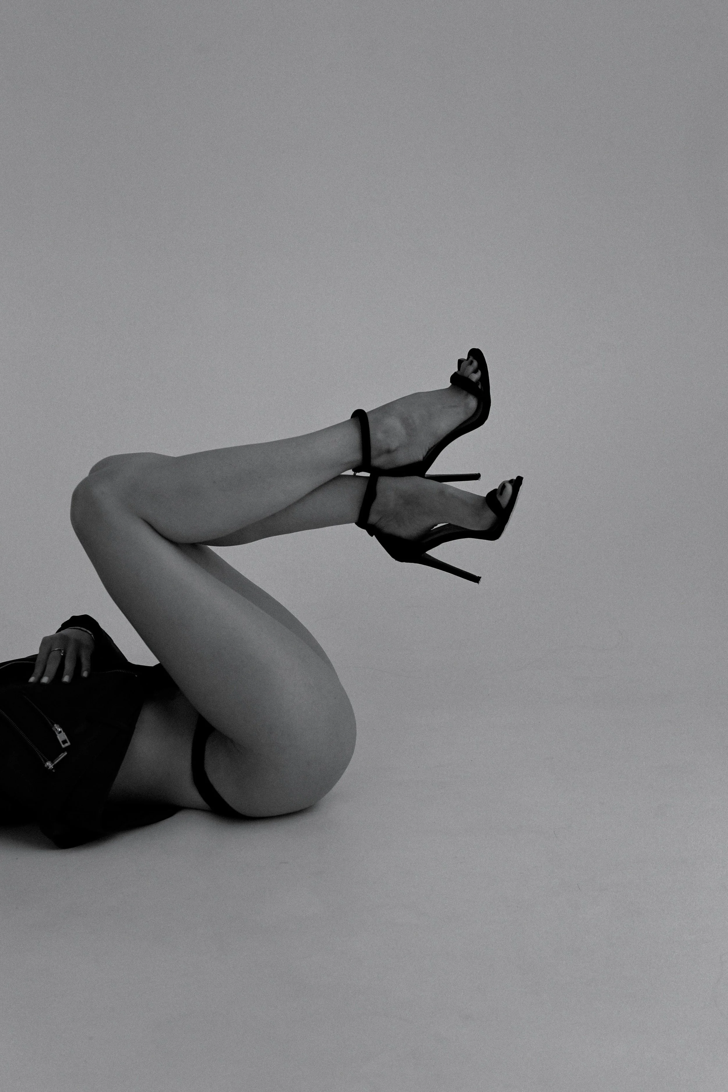 A person lying on their side wearing high-heeled sandals and black shorts, with their legs raised and crossed, in a black and white photo.