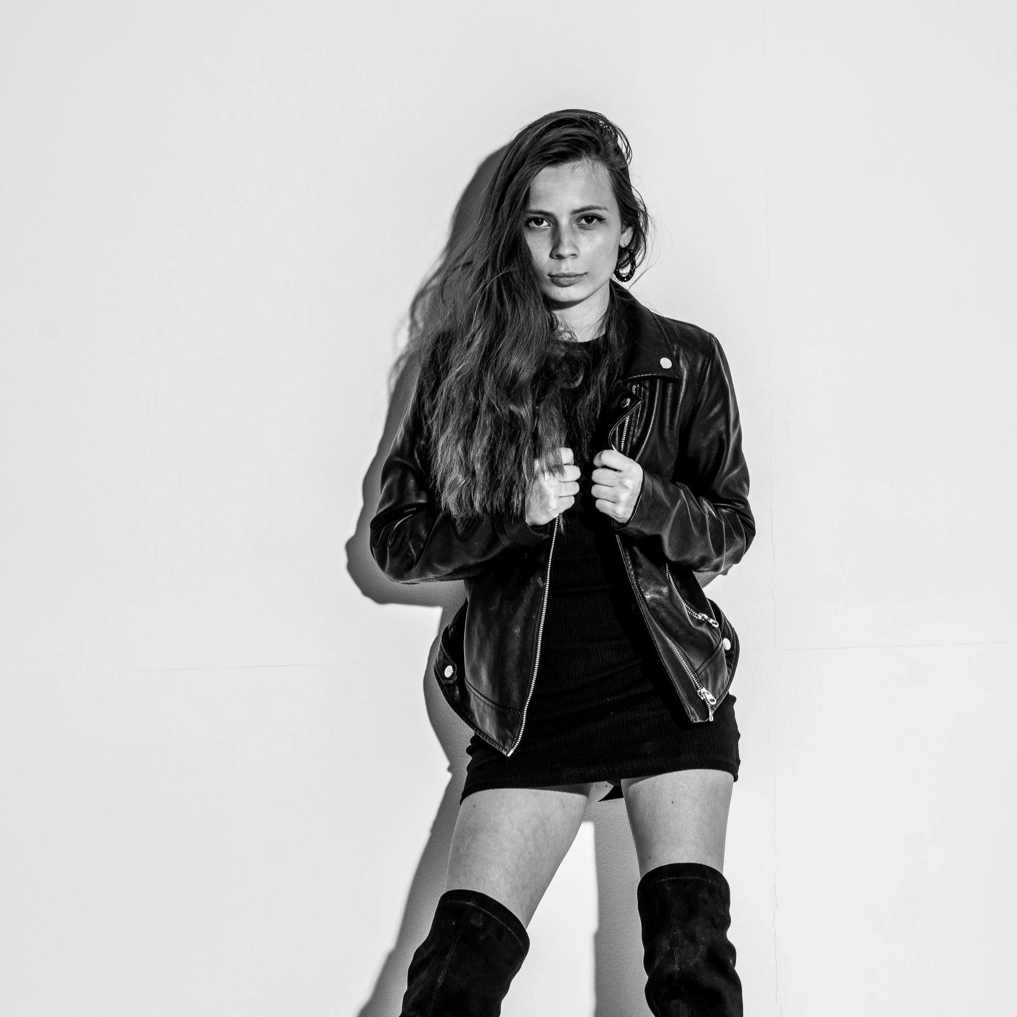 Black and white photo of a woman with long wavy hair wearing a leather jacket, a short skirt, and thigh-high boots, standing against a plain white wall.
