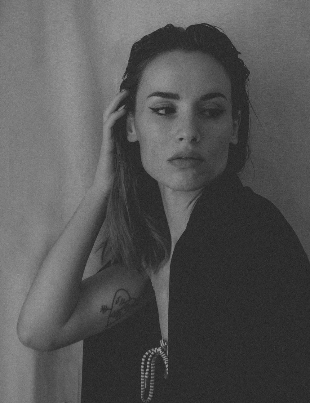 Black and white portrait of a woman with shoulder-length hair, wearing a dark top, with one hand touching her head, and a tattoo of an arrow on her upper arm.