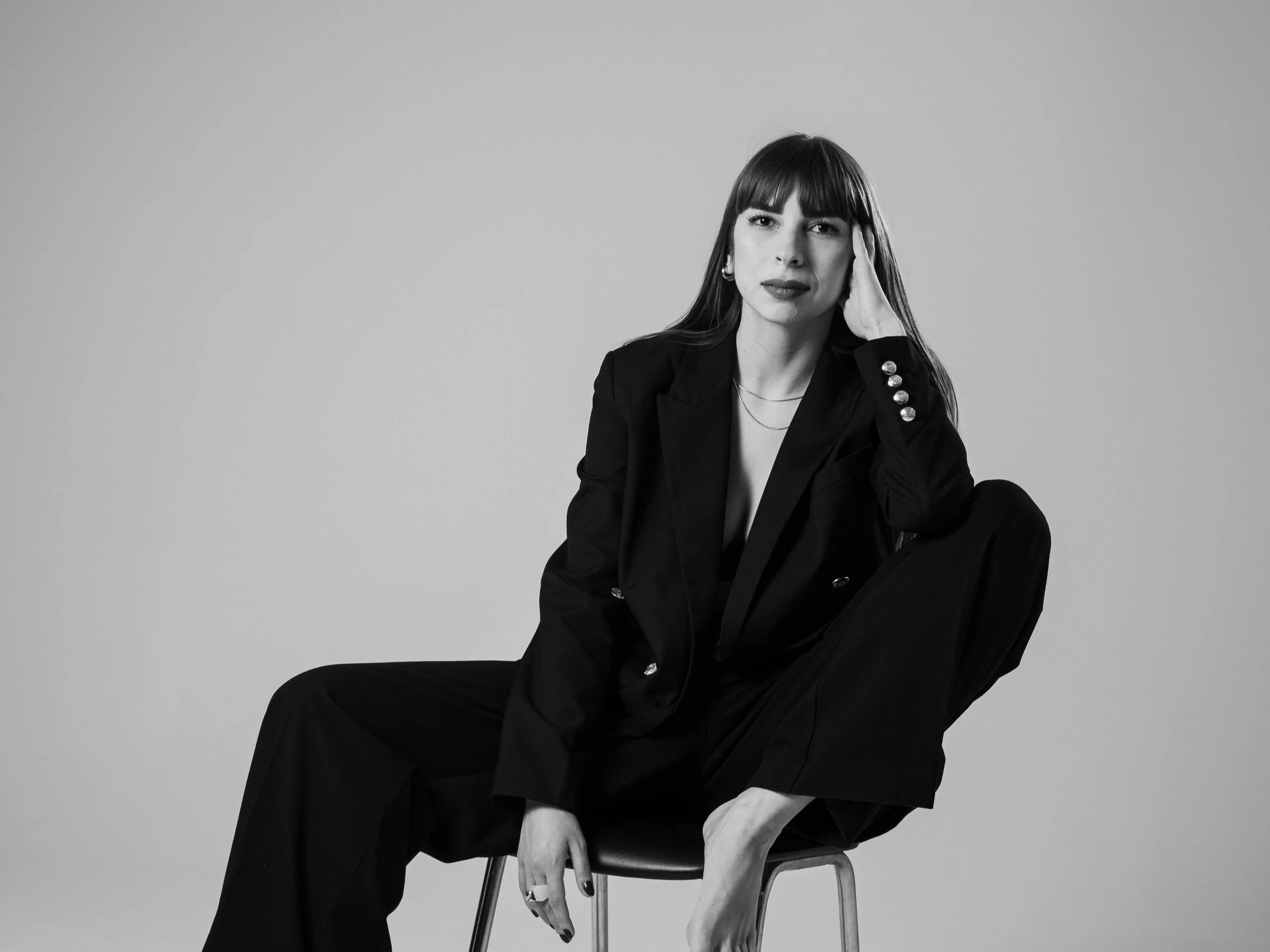A woman with long dark hair and bangs, wearing a black blazer and pants, sitting on a chair against a plain background, with one hand on her face and a serious expression.