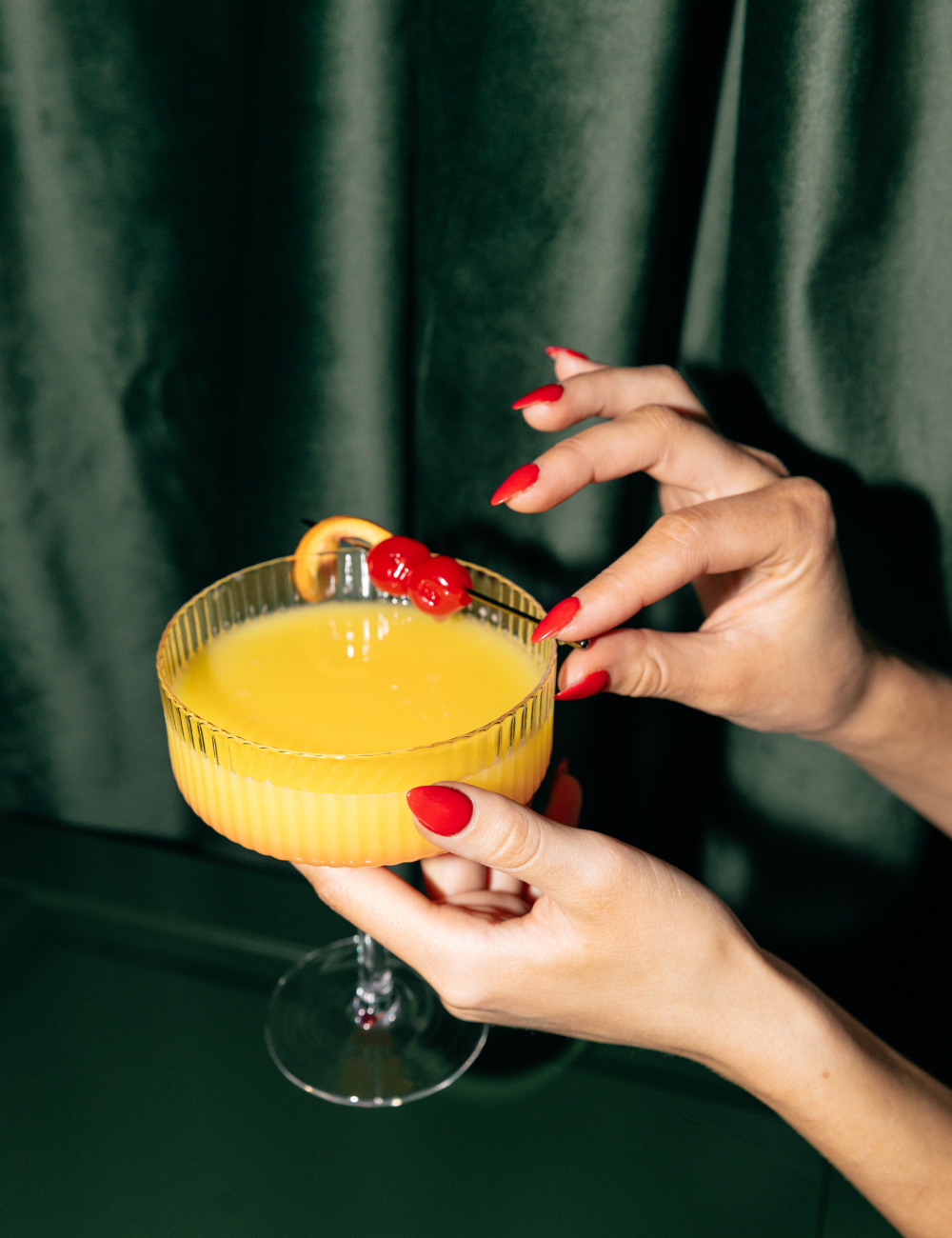 A person's hands with red nail polish holding a glass of yellow cocktail with a lemon slice and red berries garnish, against a dark green curtain background.