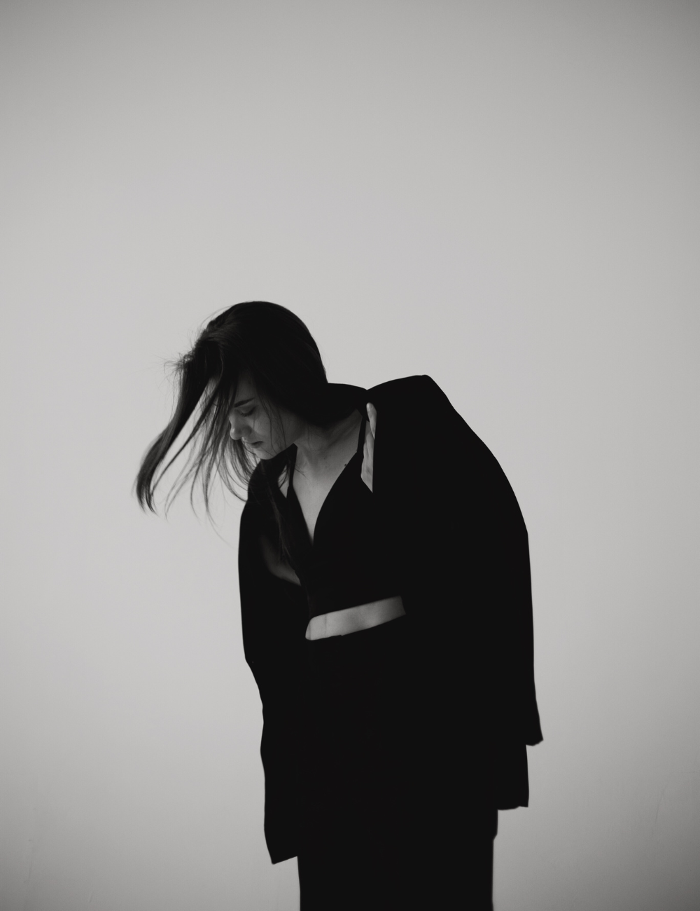 A black and white photo of a woman with dark hair, wearing a black blazer, with her head bowed and hair slightly lifted.