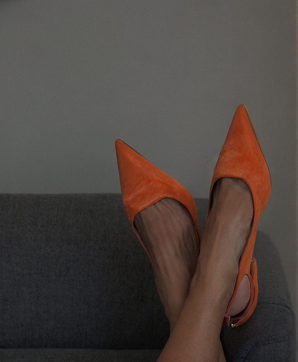 Person wearing sheer stockings and orange high heel shoes with pointed toes, sitting on a dark gray sofa.