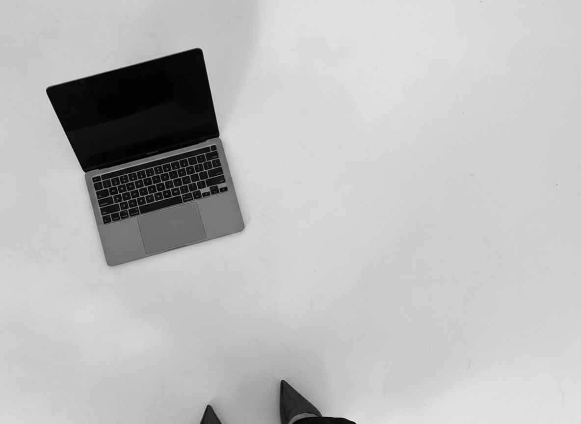 Top-down view of a laptop on a white surface with a person’s black shoe visible at the bottom of the frame.