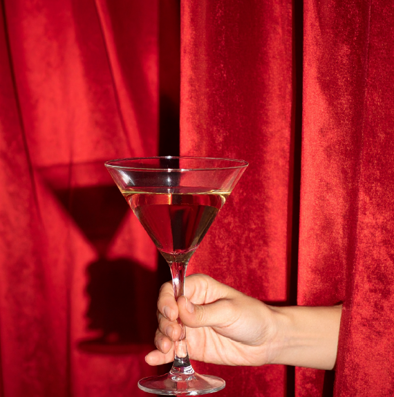 A hand holding a martini glass filled with red liquid in front of red curtains.