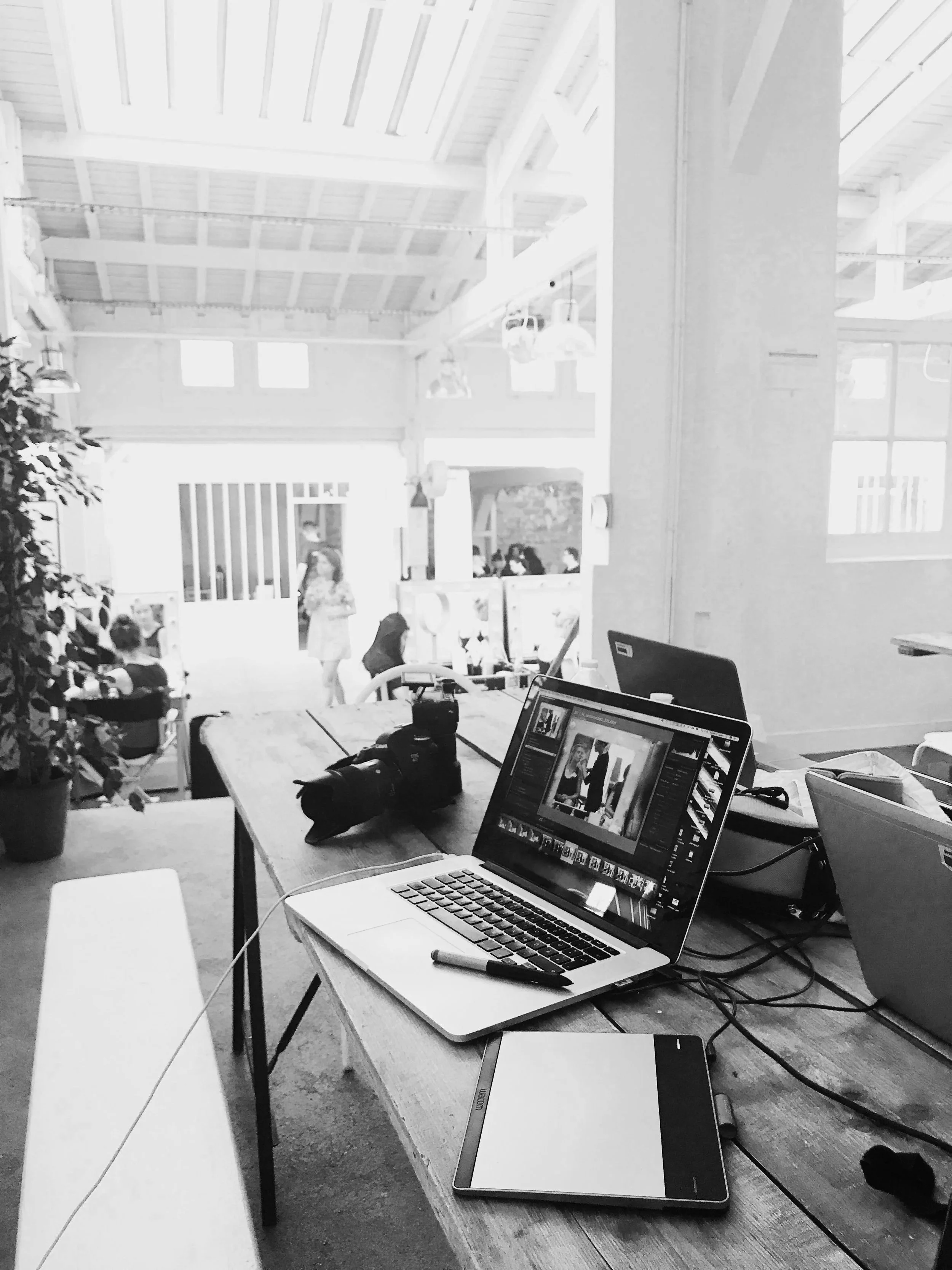 A workspace with a laptop, camera, pen, tablet, and other equipment on a wooden table in a bright, modern office with large windows and people in the background.