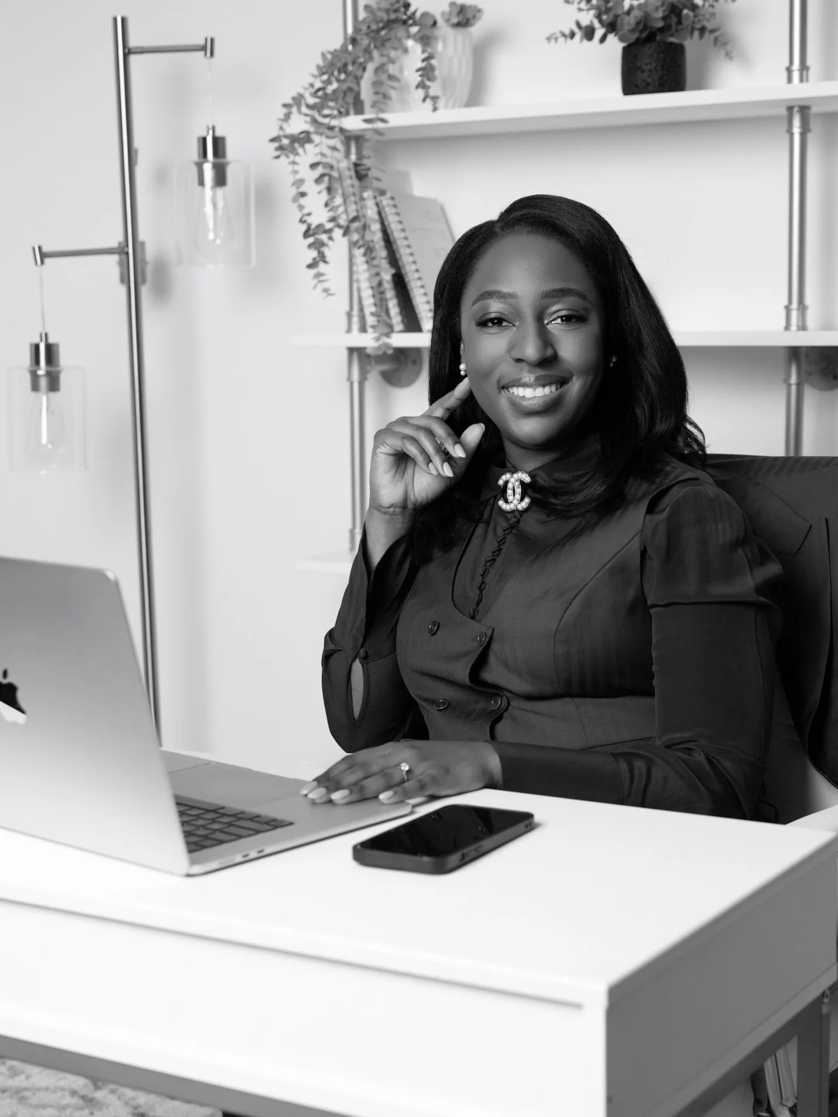 A smiling woman in business attire sitting at a desk with a laptop and smartphone, in a modern office with shelves and decorative plants in the background.