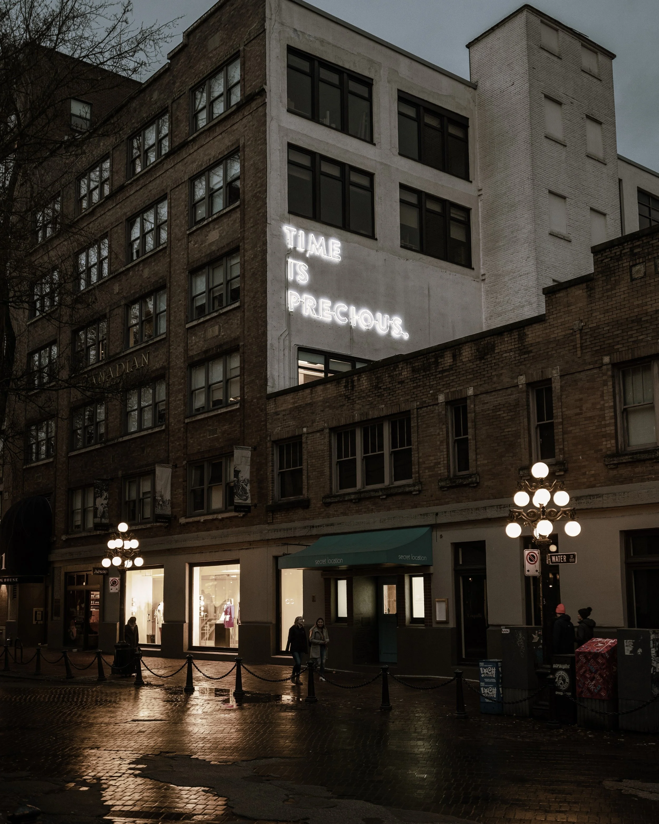 Urban street scene at dusk with a multi-story brick building featuring a white wall with neon sign reading 'TIME IS PRECIOUS.' People walking along the wet sidewalk, lit by street lamps, with retail stores on the ground floor.