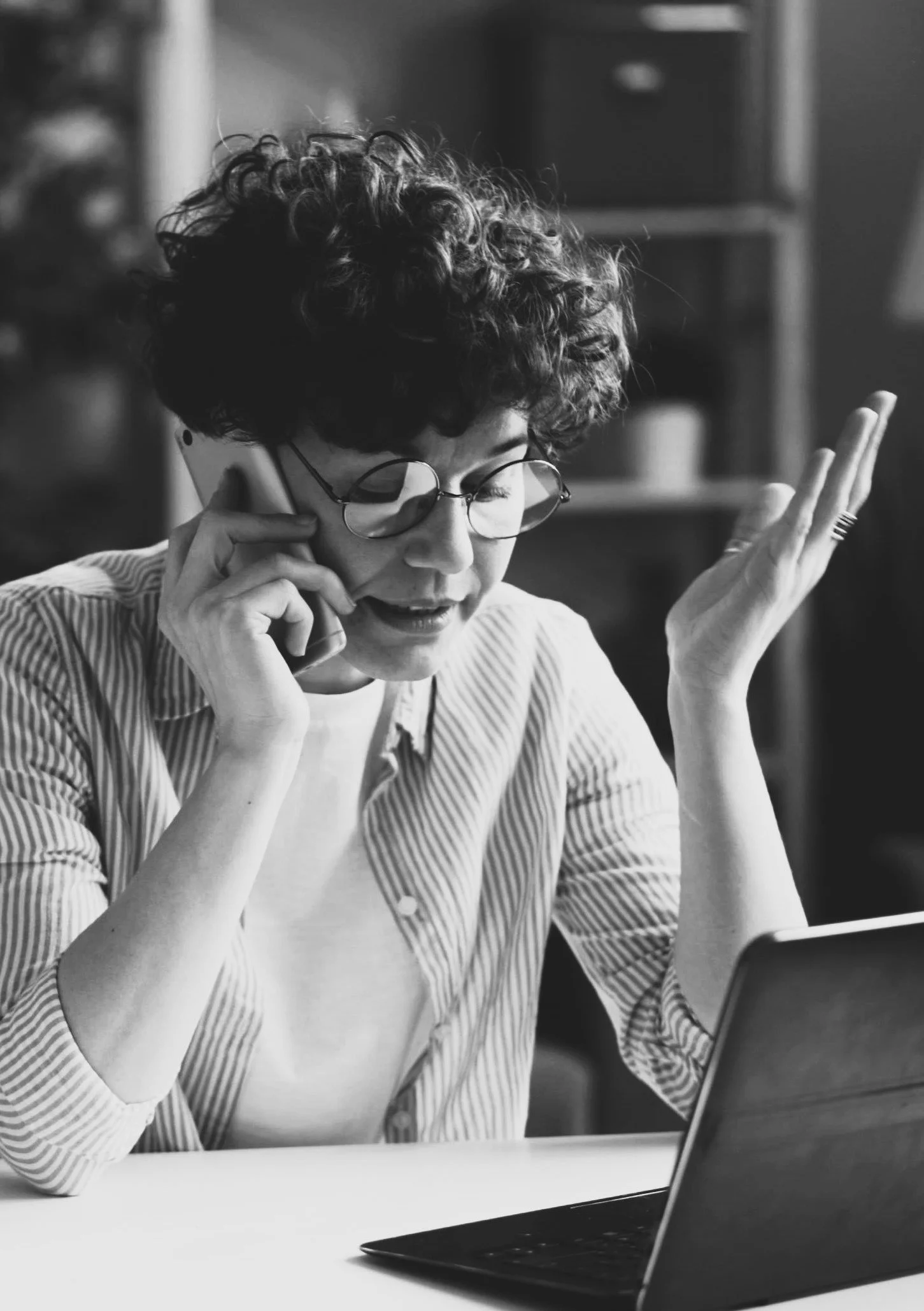 A woman with curly hair and glasses, talking on a phone while looking at a laptop, with a surprised or concerned expression.
