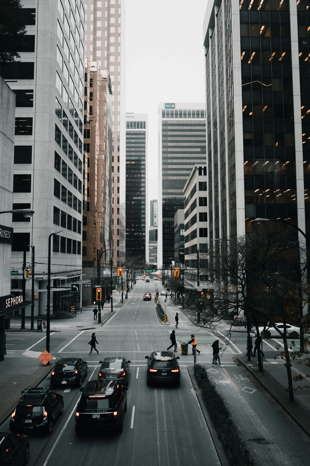 City street scene with tall office buildings, cars stopped at traffic lights, and pedestrians crossing the street in a downtown area.