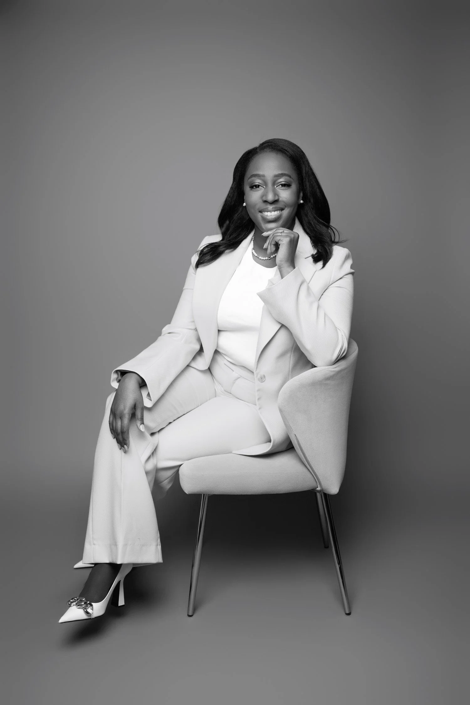 Black and white portrait of Thelma Zindoga sitting on a chair, smiling, dressed in a light-colored suit and high heels, against a plain background.