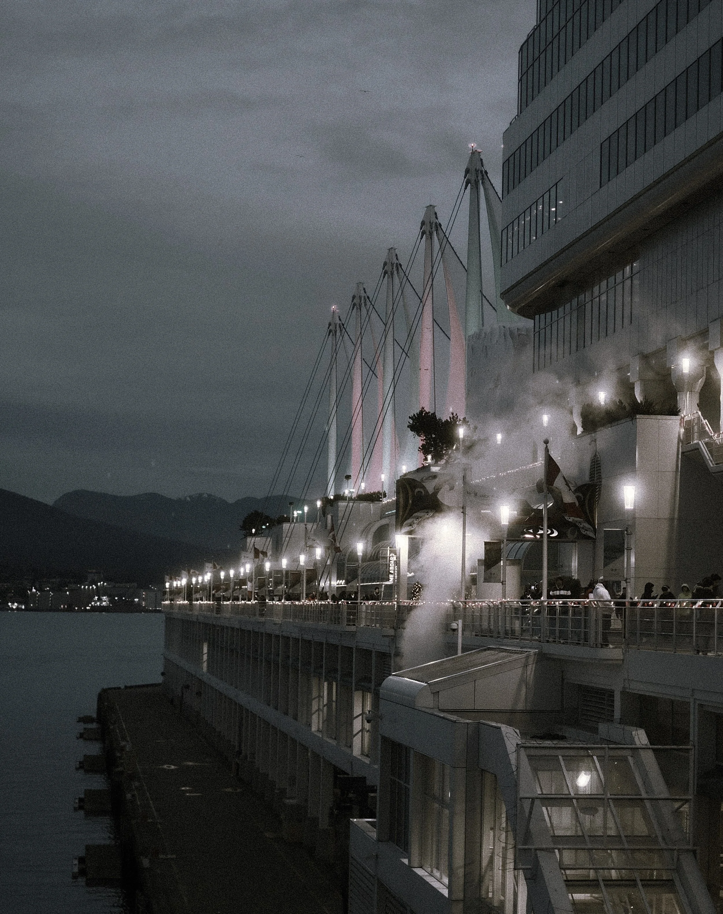 Nighttime scene of a boat or ship docked at a pier with steam rising, illuminated by many lights, with mountains and water in the background.