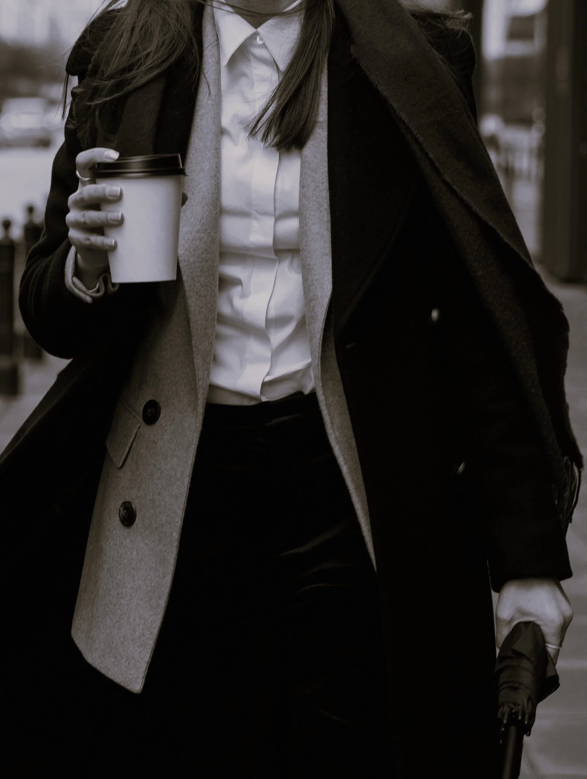 Close-up of a woman wearing a white shirt, gray blazer, and black coat, holding a coffee cup in her left hand and an umbrella in her right hand, walking outdoors.