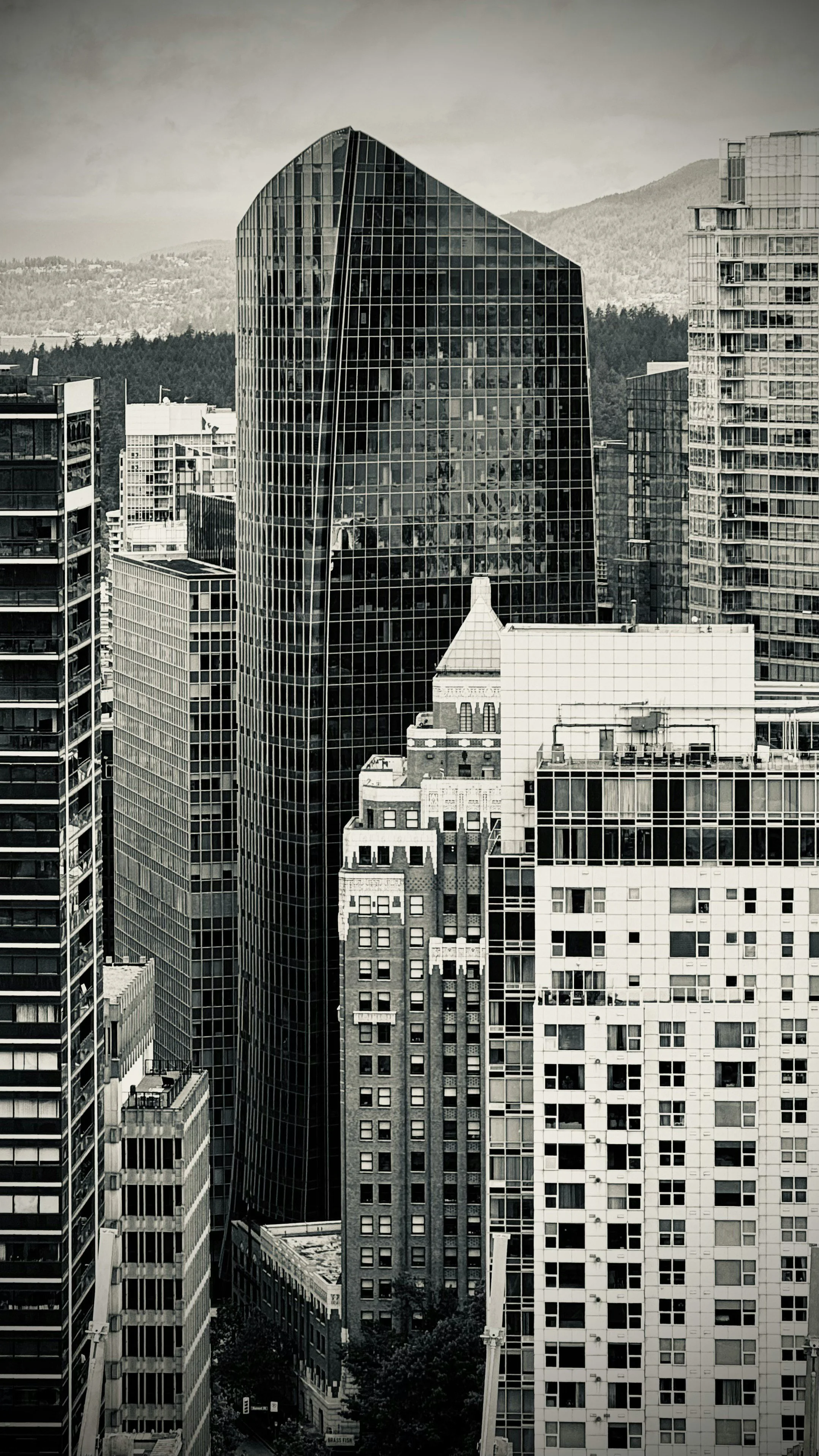 Black and white photo of urban cityscape with tall buildings, including a prominent glass skyscraper, with mountains in the background.