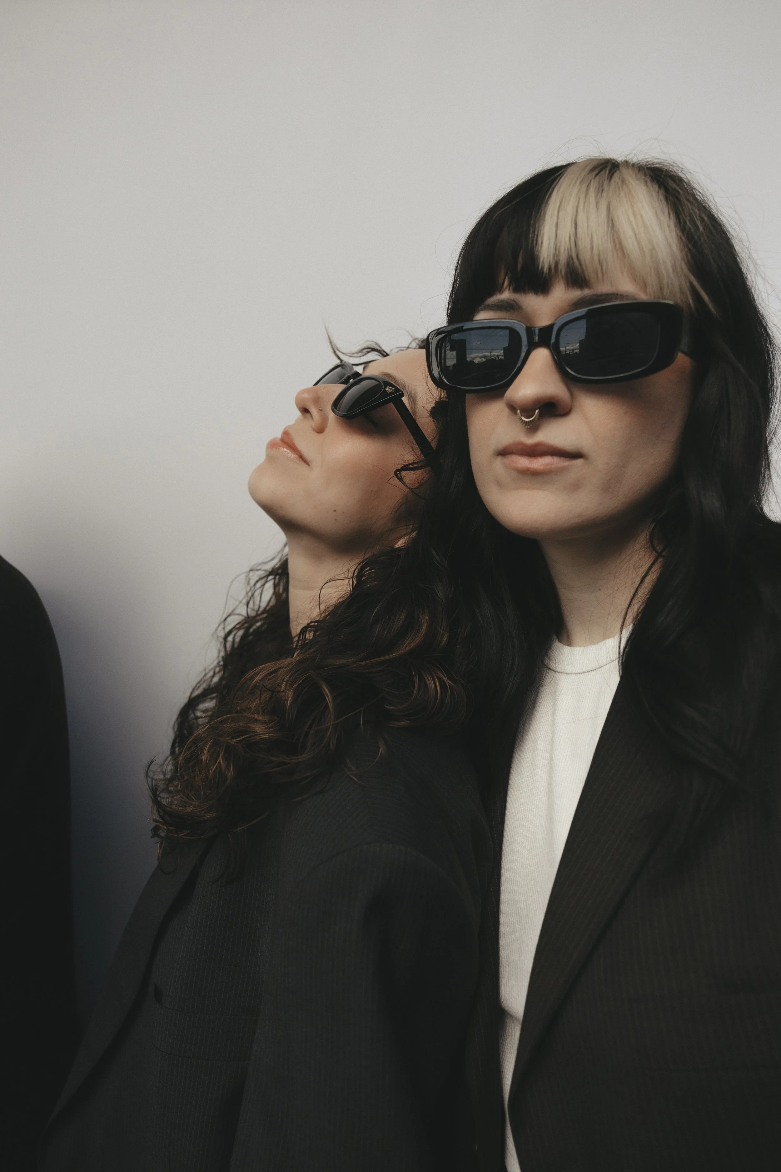 Two women with dark sunglasses and black hair, one with a blonde streak, dressed in black jackets, standing against a plain light background.
